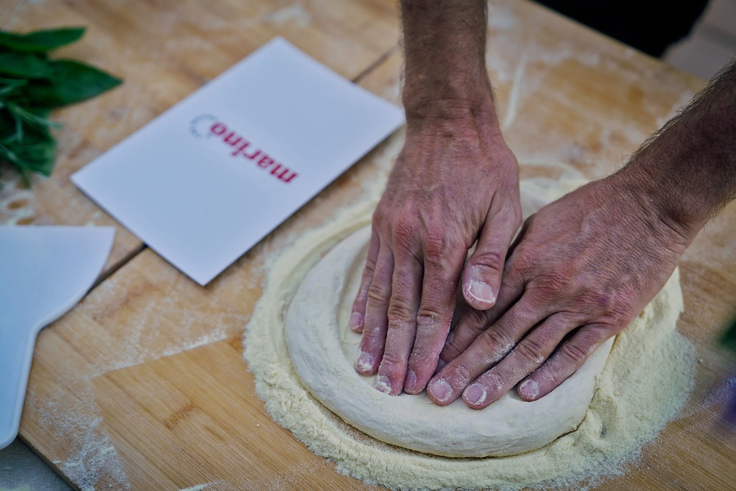 Close-up of two hands gently pressing the edge of a pizza dough ball on a semolina-dusted wooden board, with a Marino & The Dough masterclass booklet tucked beside it.
