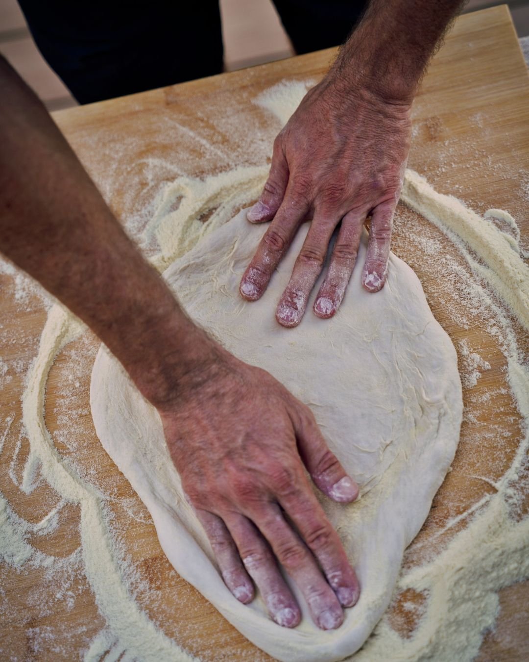 Flour-dusted hands shape pizza dough on a wooden bench during a Marino & The Dough masterclass, surrounded by semolina and signs of hands-on craft.