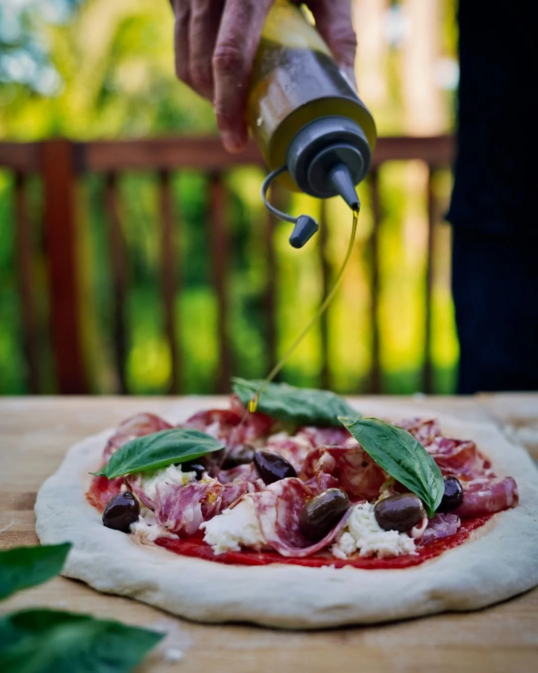 Olive oil being drizzled over a freshly topped Neapolitan style pizza with basil, olives and cured meat before firing during a Marino & The Dough masterclass