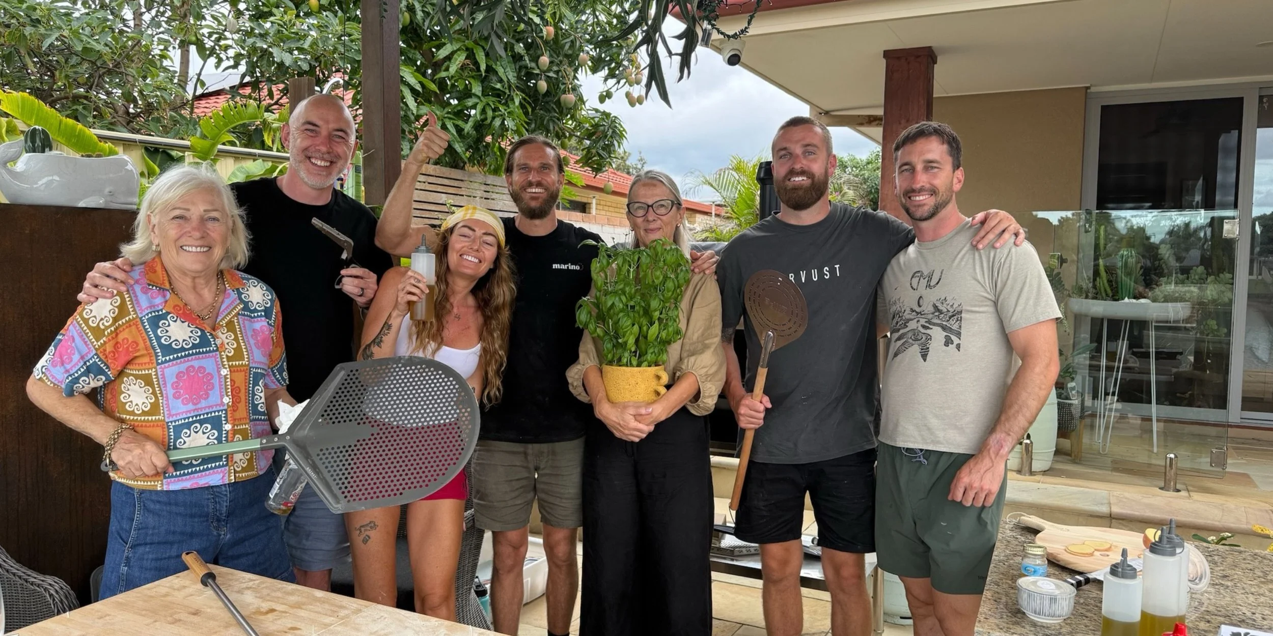Group of guests and Luke Marino smiling outdoors with pizza peels and fresh basil during a private Marino & The Dough masterclass on the Gold Coast