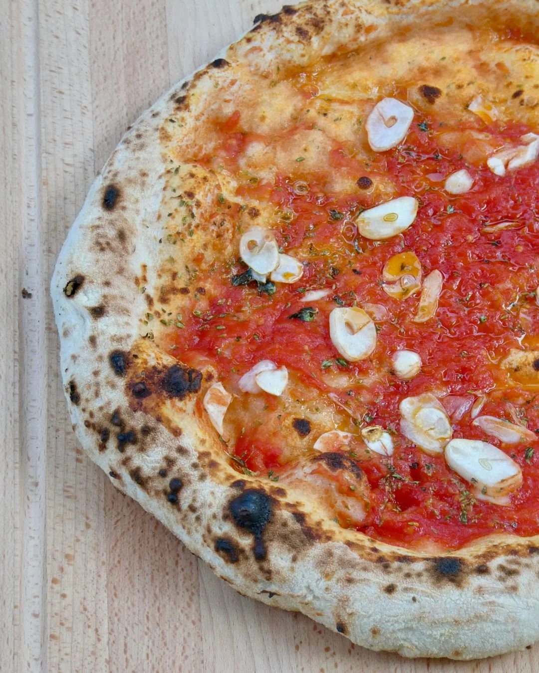 Close up of a marinara pizza with bright tomato sauce, sliced garlic, herbs and olive oil on a blistered wood fired crust, served on a pale timber board.