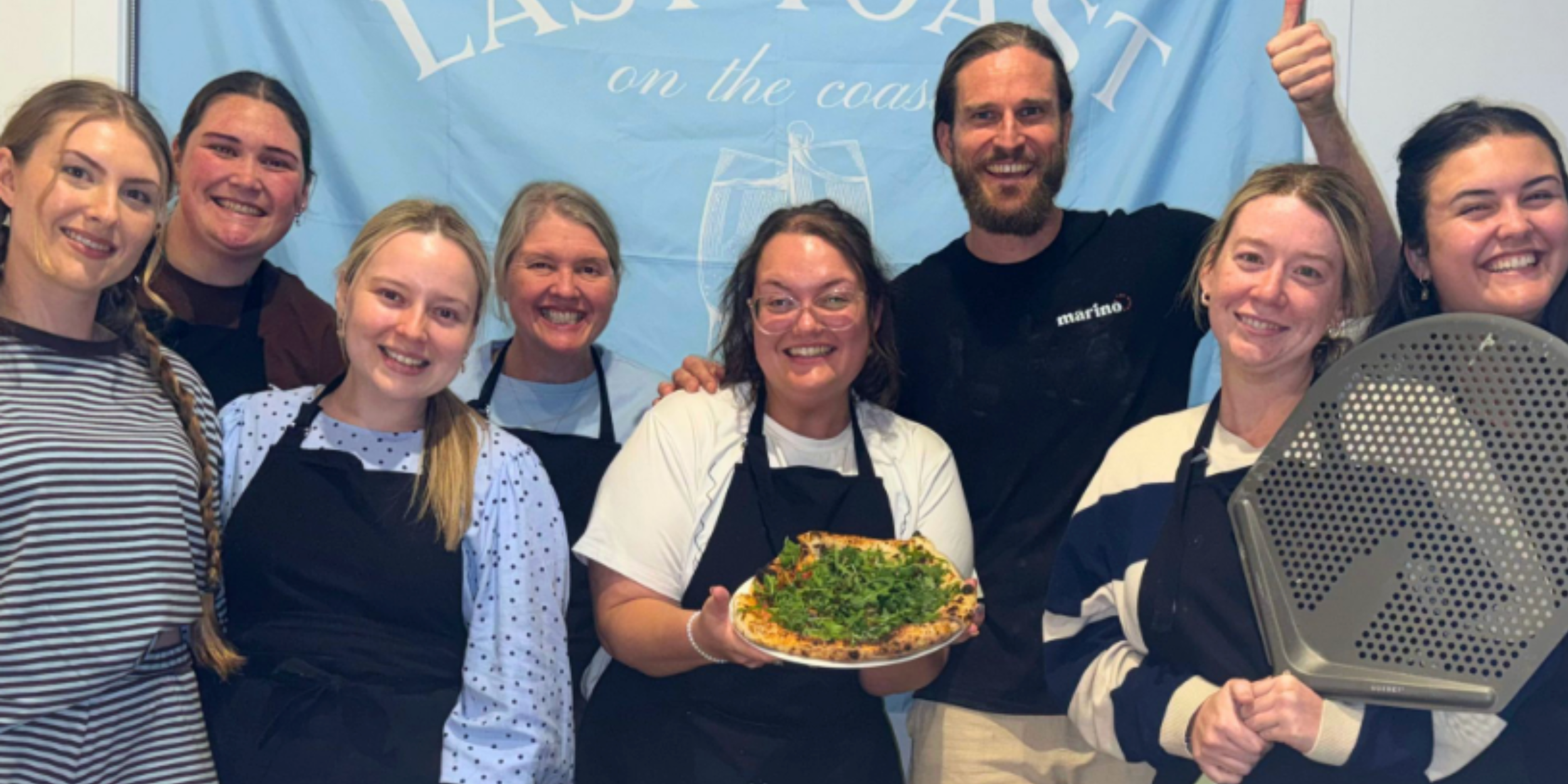 Hen’s party group in aprons smiling with their pizza instructor, with one guest holding a finished pizza and another holding a pizza peel.