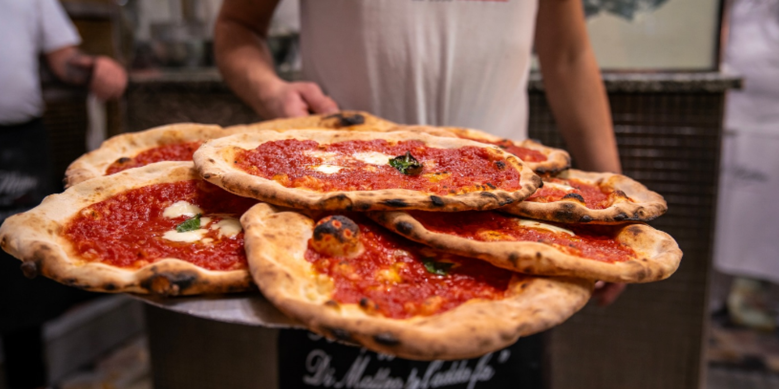 Pizzaiolo carrying a large tray stacked with freshly baked Neapolitan pizzas topped with tomato, mozzarella and basil.