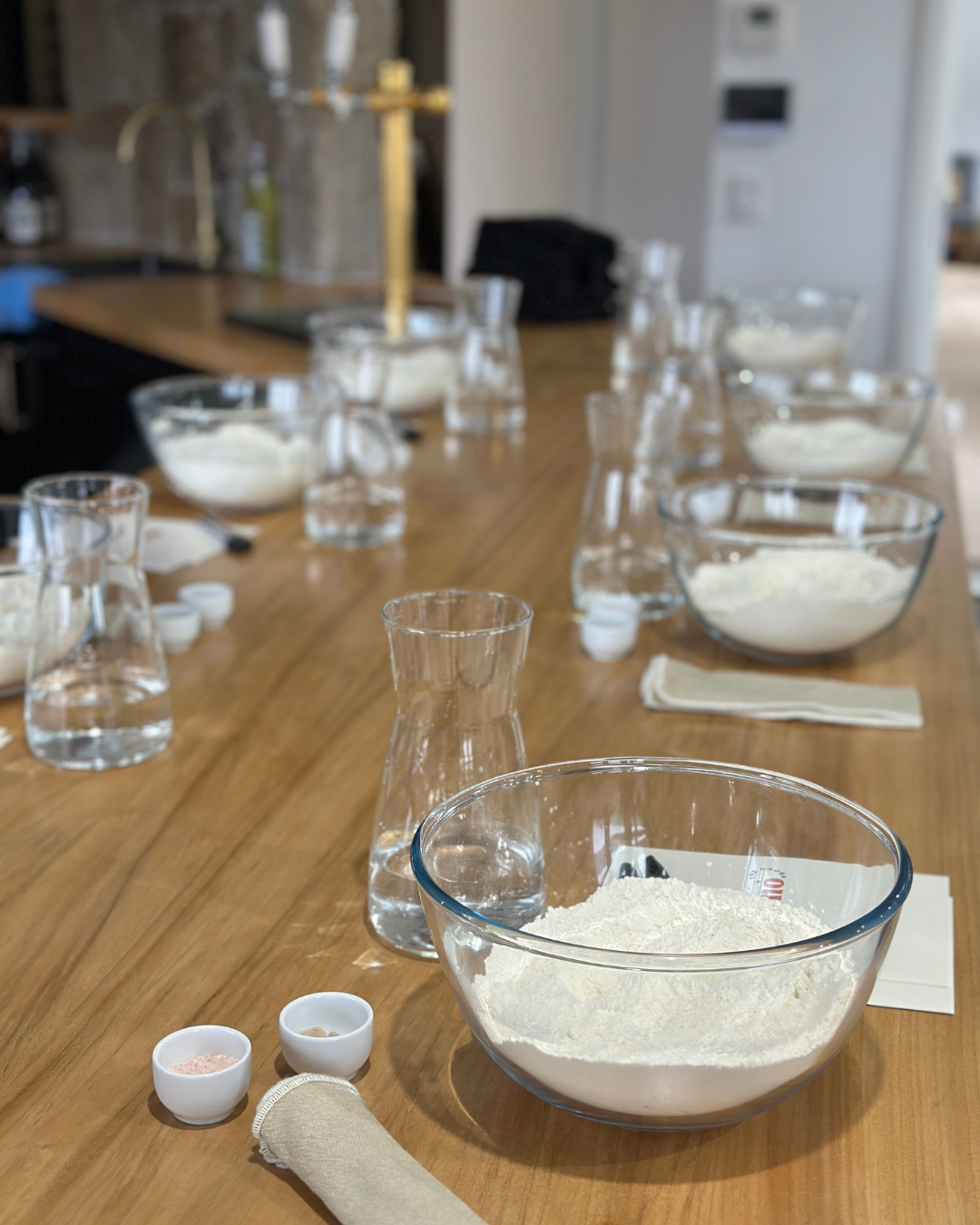Glass bowls of flour, water jugs, and salt set up for a hands-on pizza dough masterclass by Marino & The Dough, displayed on a timber kitchen bench.