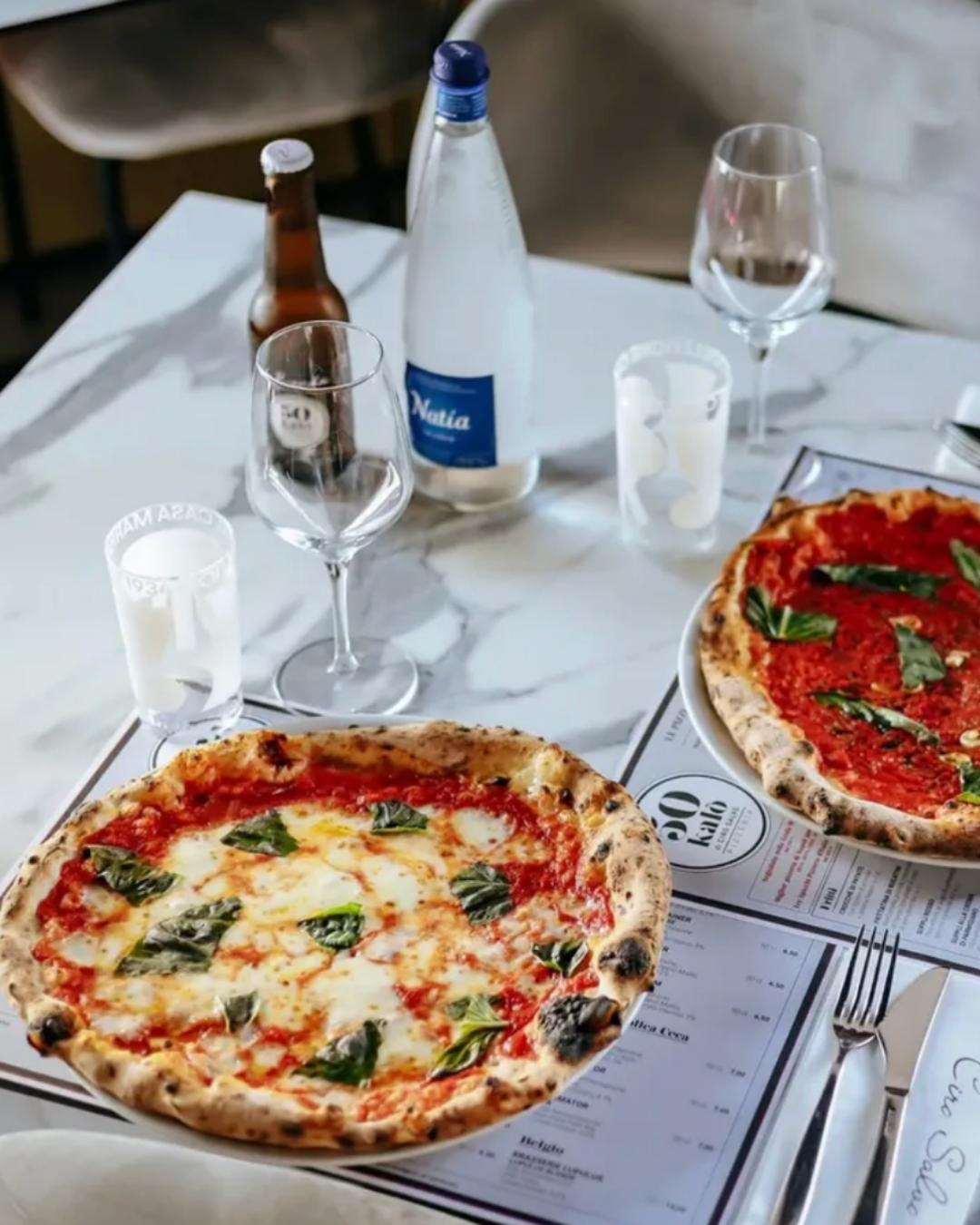 Two Neapolitan pizzas with blistered crusts, tomato, mozzarella and fresh basil served on a marble table with glassware and bottled drinks.