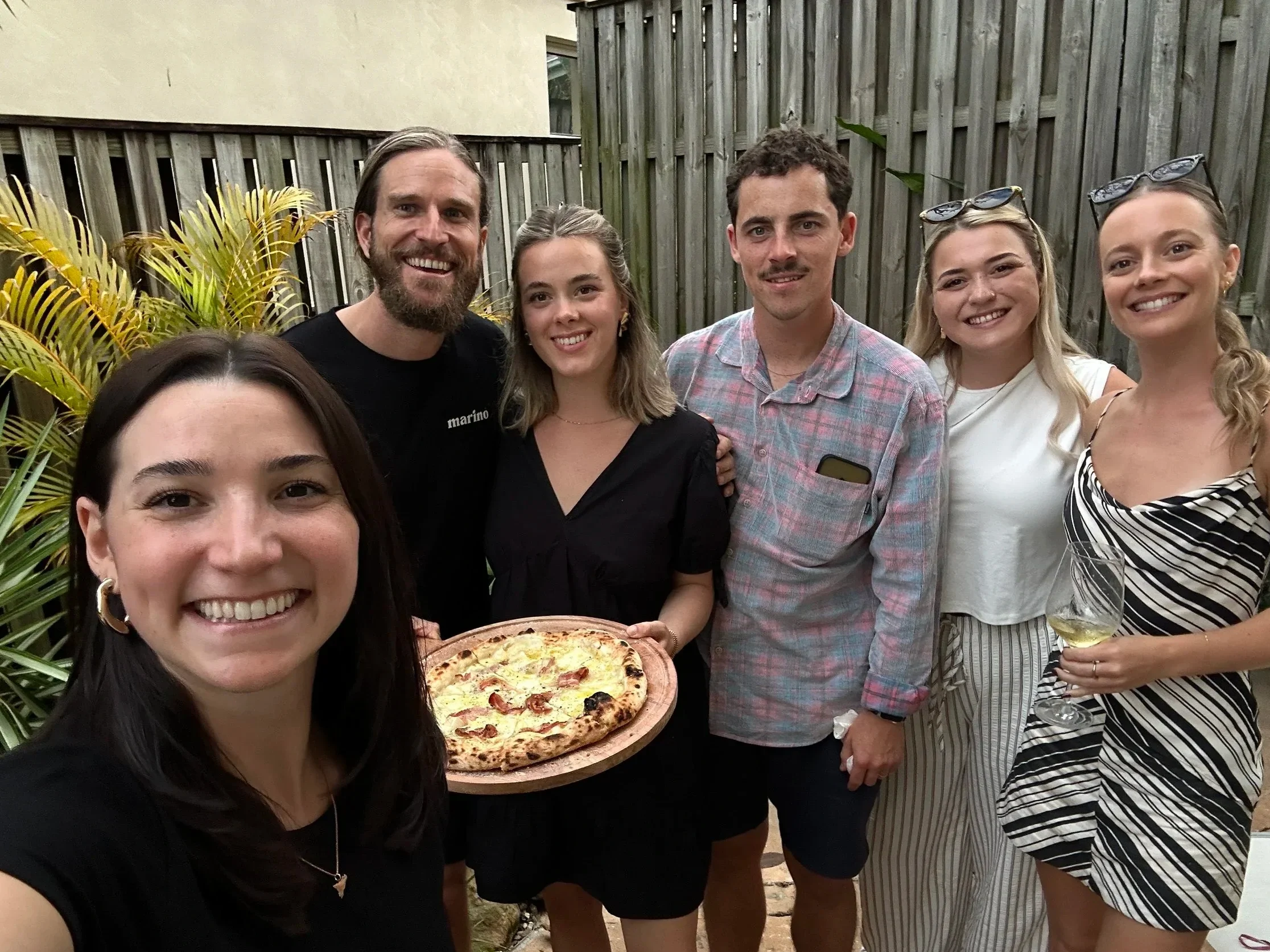 Group selfie after a pizza masterclass, smiling outdoors and holding a freshly baked pizza and wine