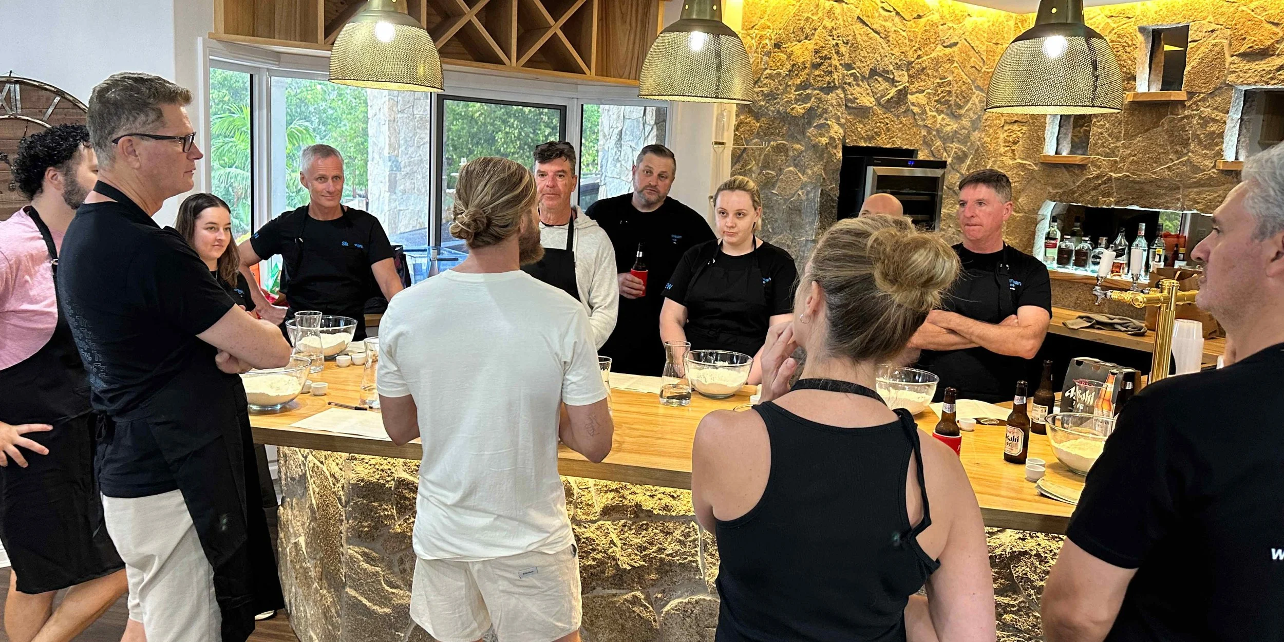 Smiling team posing after a Marino & The Dough pizza masterclass, wearing aprons and standing together at a scenic outdoor venue surrounded by greenery.