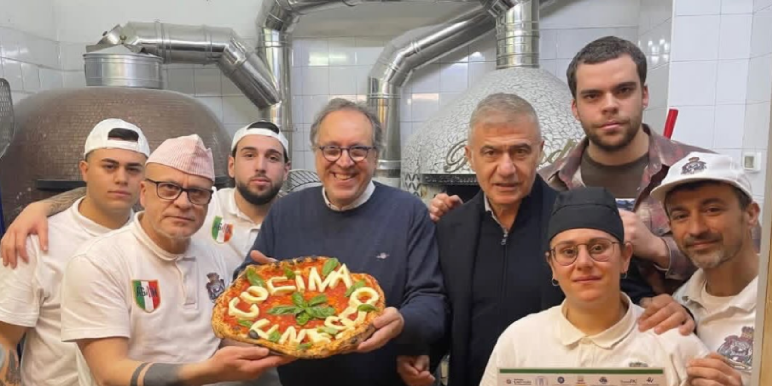 Group of chefs and supporters in a pizzeria kitchen holding a basil-topped pizza and a sign celebrating an Italian cuisine UNESCO victory.