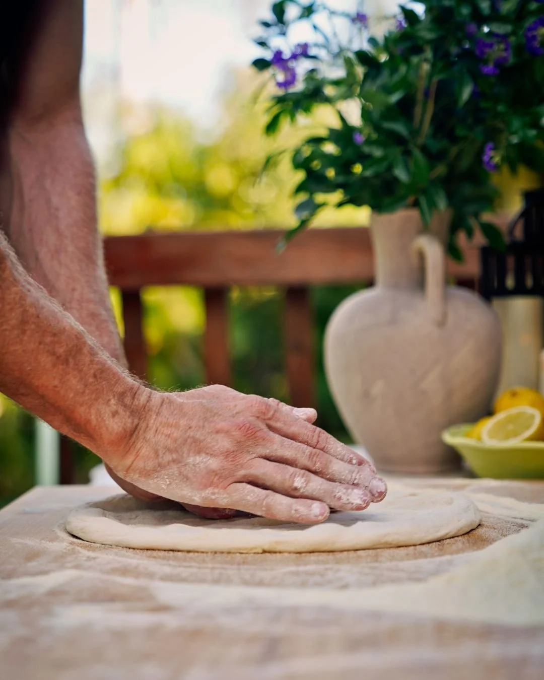 A floured hand shapes pizza dough on a rustic wooden bench in an outdoor Marino & The Dough masterclass, with soft sunlight, lemons, and a clay vase in the background.
