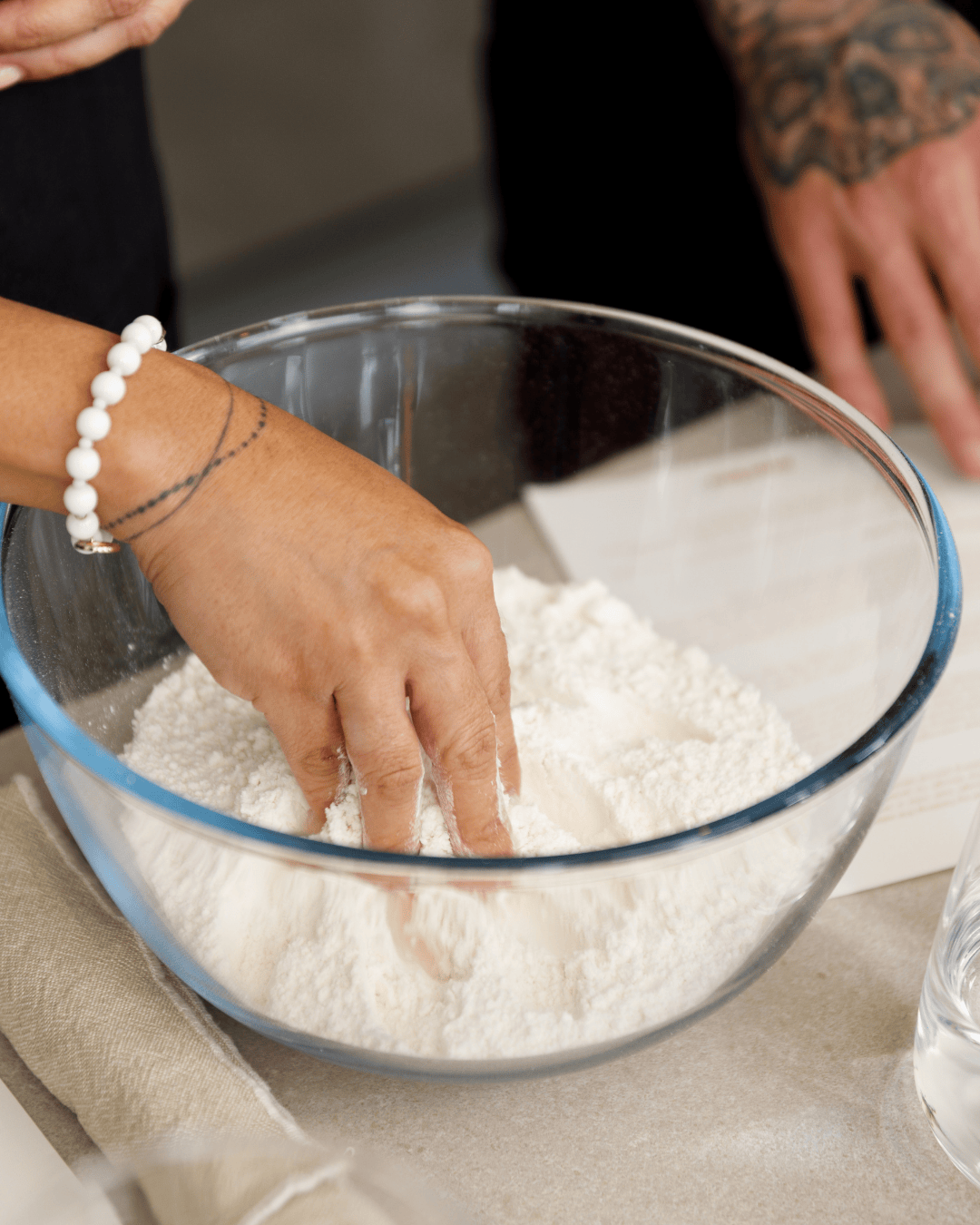 Close-up of hands mixing flour in a glass bowl during a pizza masterclass, showing the early stages of dough preparation with measured ingredients beside the workspace.
