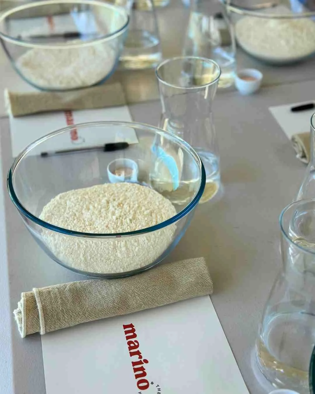 A close-up of individual dough prep stations at a Marino & The Dough masterclass, featuring bowls of flour, water carafes, salt, and printed recipe guides on a clean table setting.