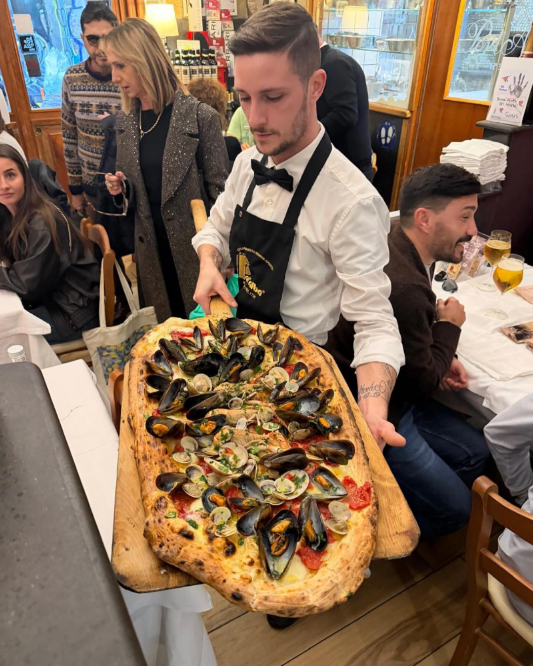 Restaurant waiter carrying an oversized seafood pizza topped with mussels and clams through a busy dining room.