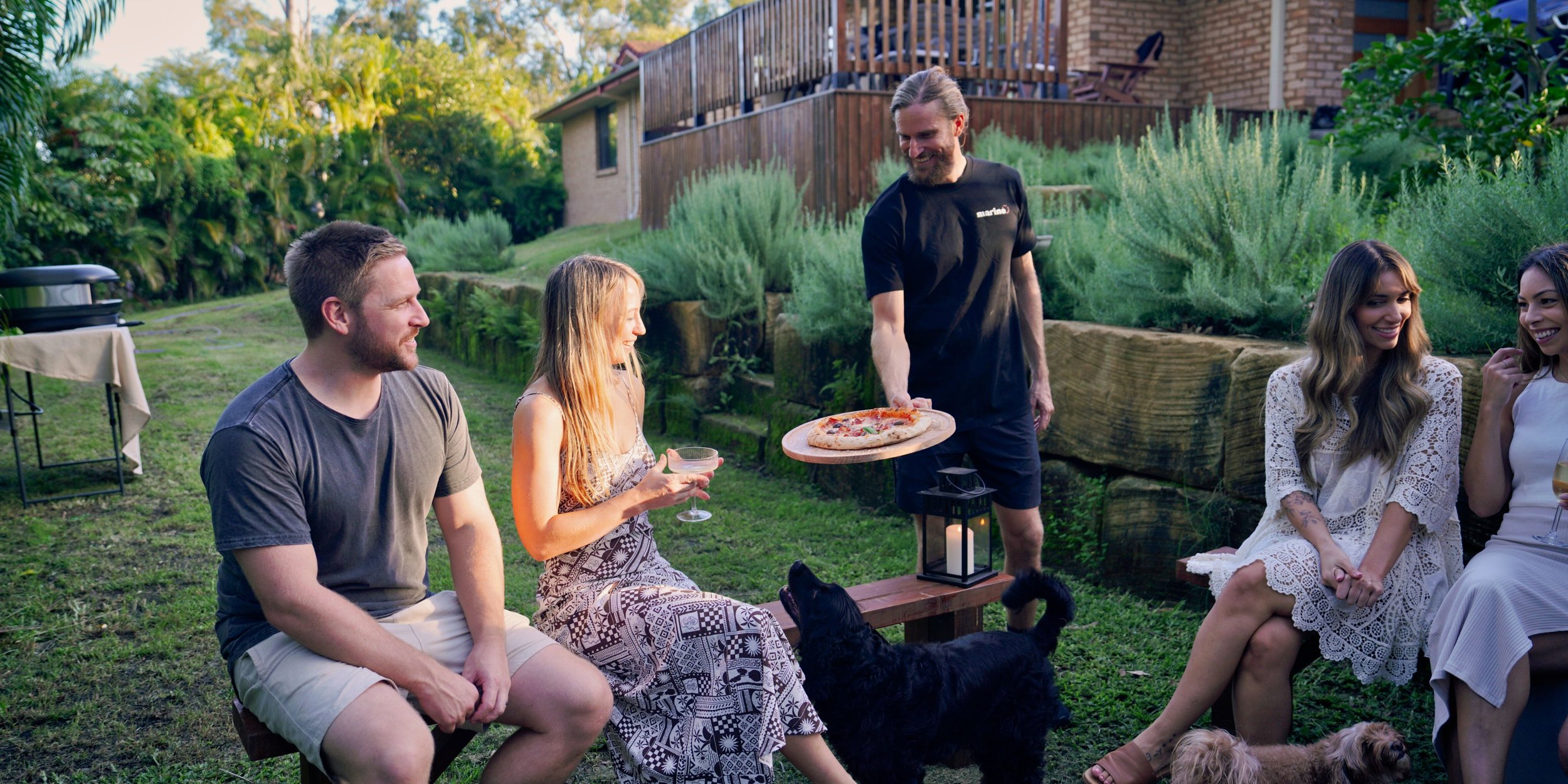 Luke Marino serving a wood-fired pizza to guests at a backyard masterclass on the Gold Coast. Relaxed, social vibe with drinks, pets, and friends enjoying the afternoon together.
