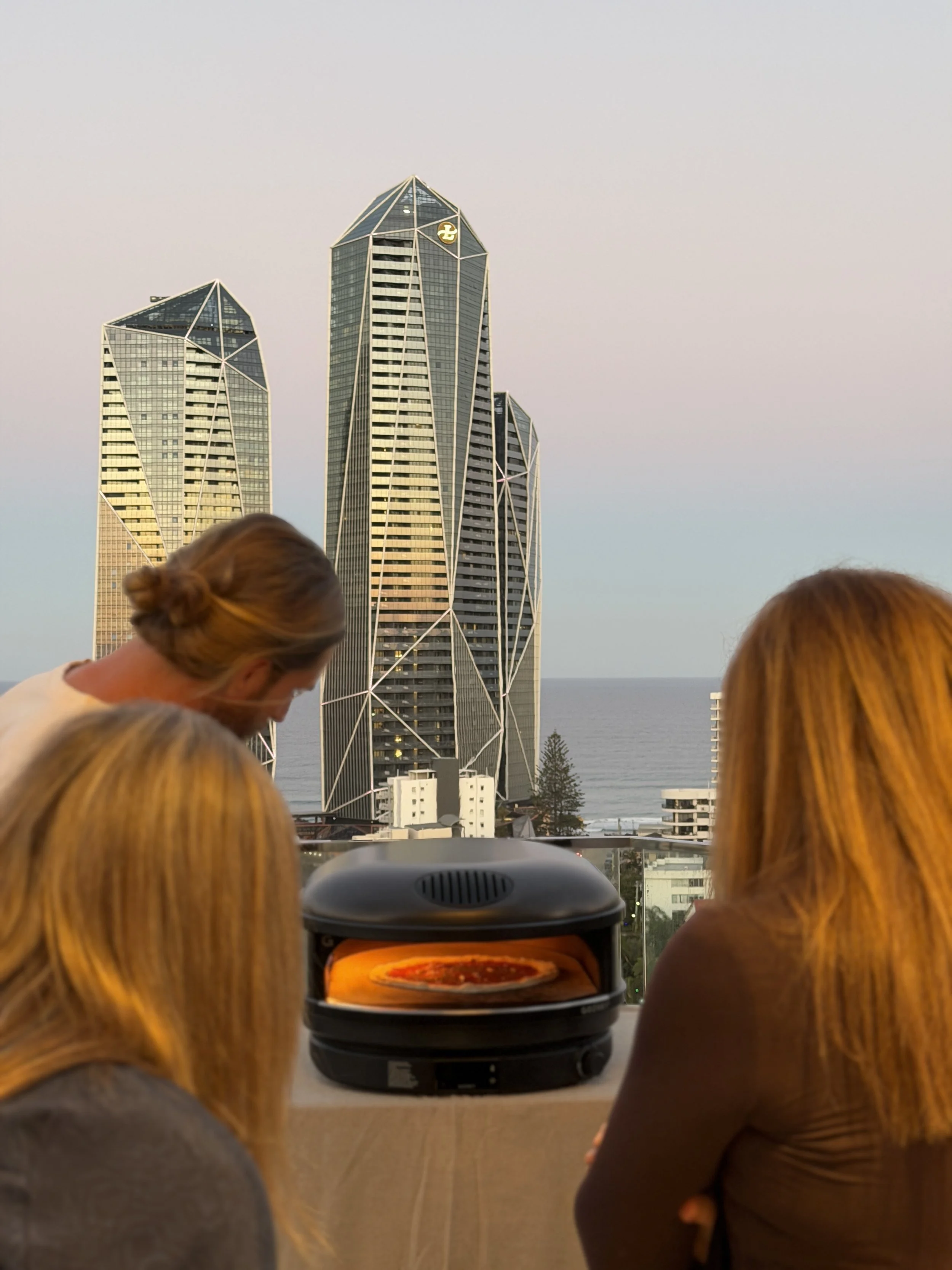 Guests watch a pizza cook in a portable oven during a Marino & The Dough masterclass on a Gold Coast balcony overlooking the city and ocean.