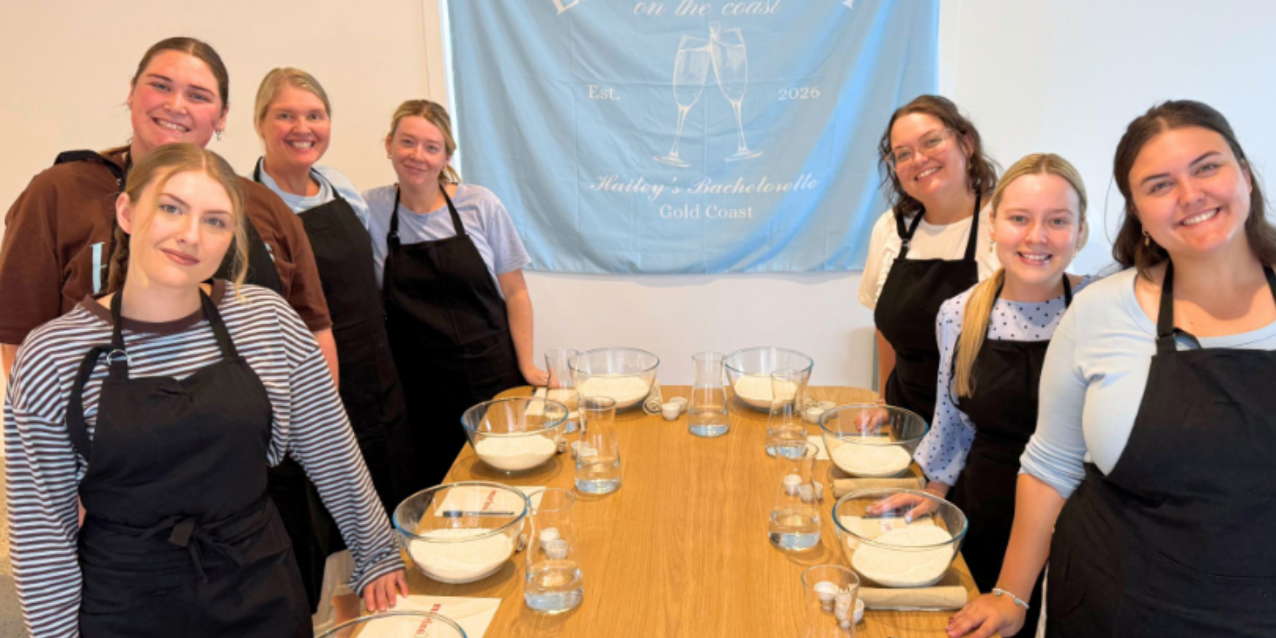 Hen’s party group wearing aprons and smiling around a timber table prepared with flour, water and pizza making equipment beneath a personalised celebration banner.