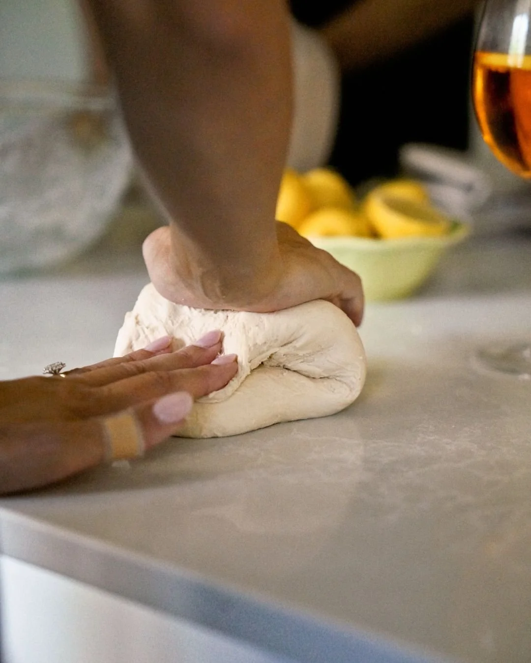 Two hands work together to knead pizza dough on a floured countertop during a Marino & The Dough masterclass, with fresh lemons and an amber drink in the soft-focus background.