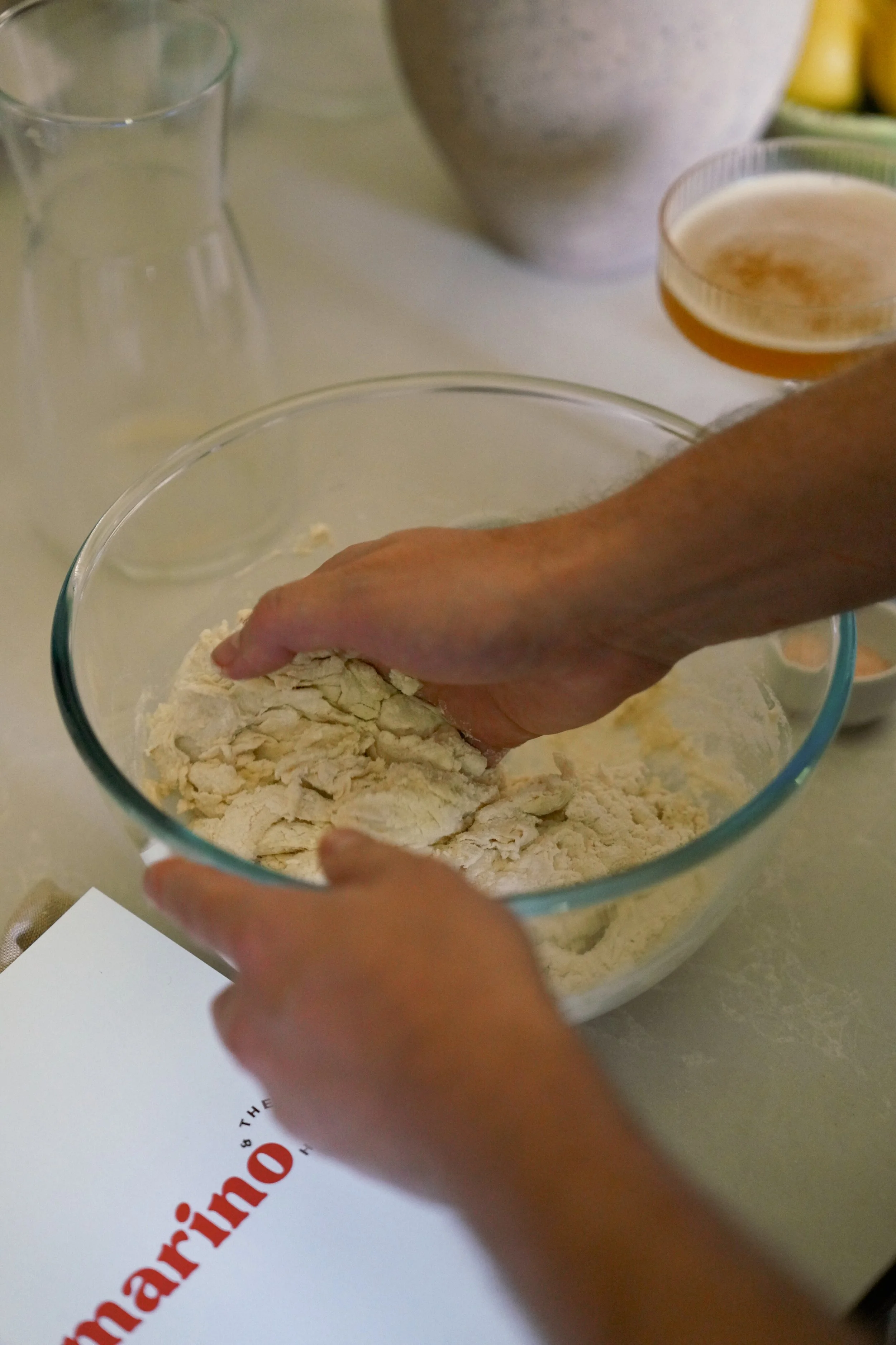 Close-up of two hands folding pizza dough in a clear glass bowl beside a Marino & The Dough masterclass booklet on a light kitchen countertop.