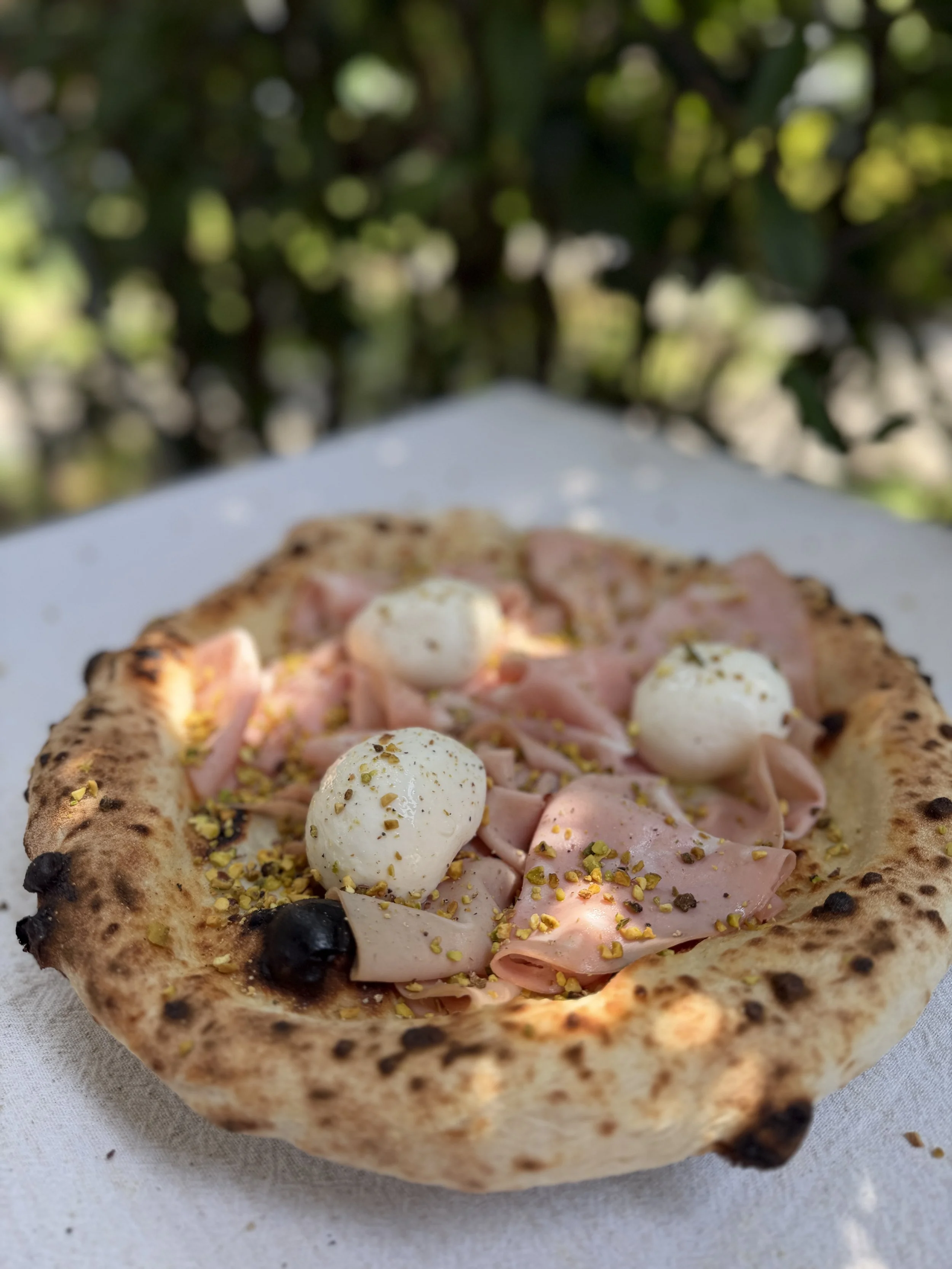 Neapolitan-style pizza topped with sliced mortadella, creamy burrata, olives, pistachios, and chili flakes on a charred crust
