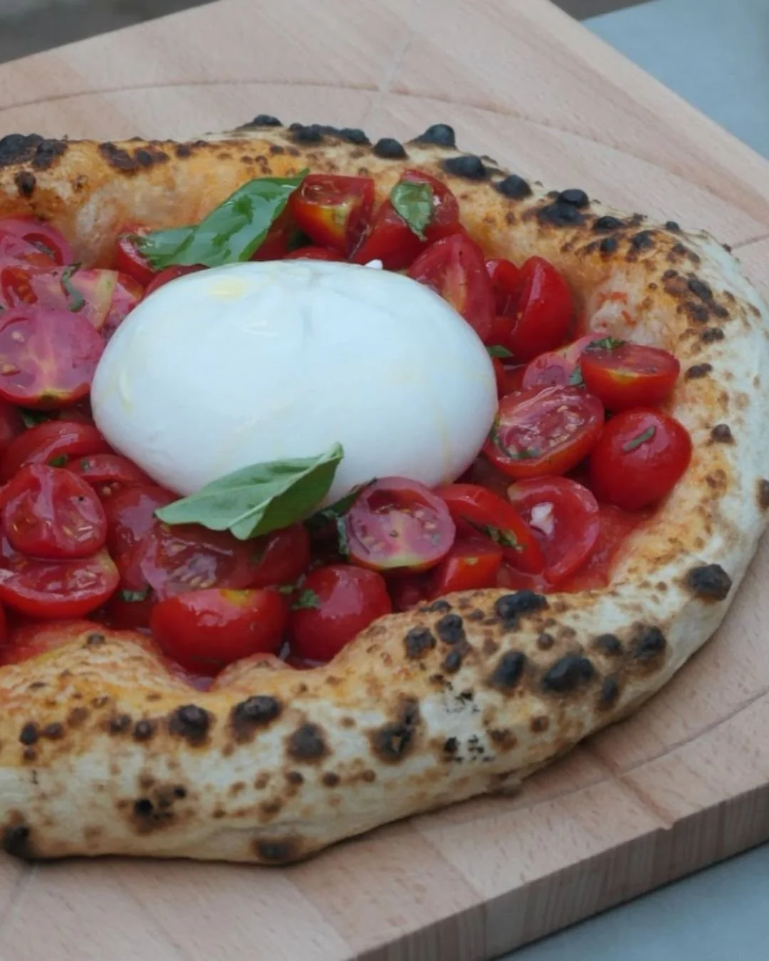 Neapolitan style pizza with blistered crust topped with fresh burrata, cherry tomatoes and basil on a wooden board during a Marino & The Dough masterclass