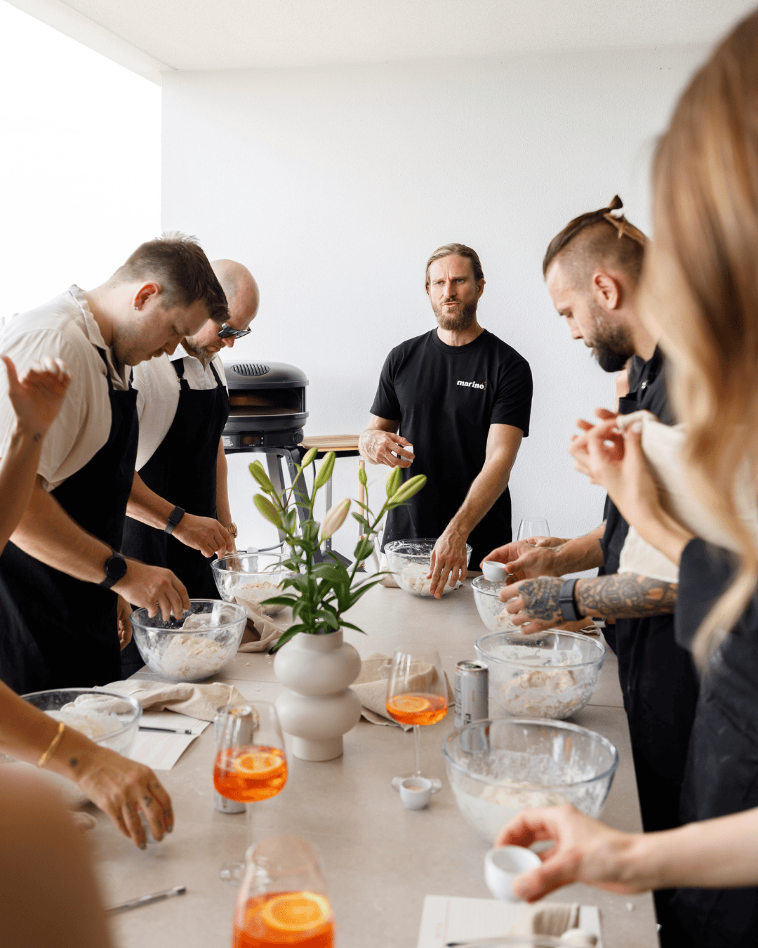 Guests gathered around a table learning to mix and shape pizza dough under guidance, with bowls of dough, wine glasses, and a relaxed home setup creating a shared cooking experience.