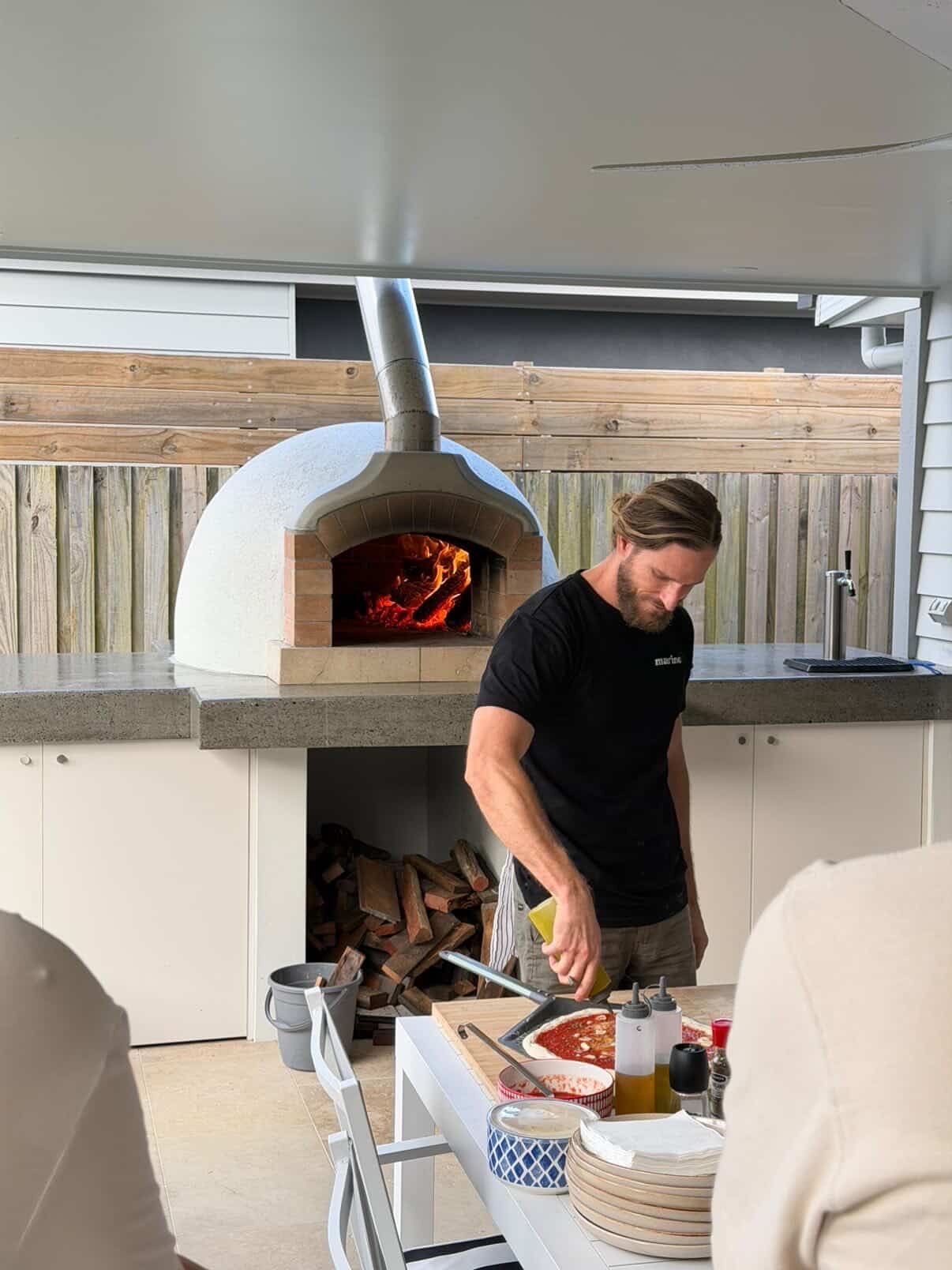 Marino & The Dough host preparing a fresh pizza with sauce and toppings beside a glowing wood-fired oven at an outdoor masterclass
