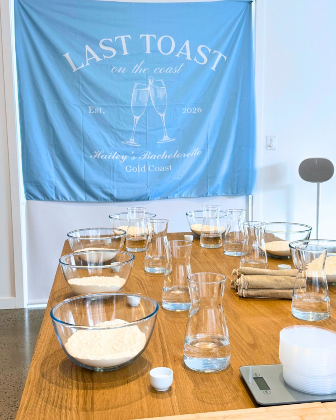 Long timber table prepared with flour, water and pizza making equipment beneath a personalised blue hen’s party banner.