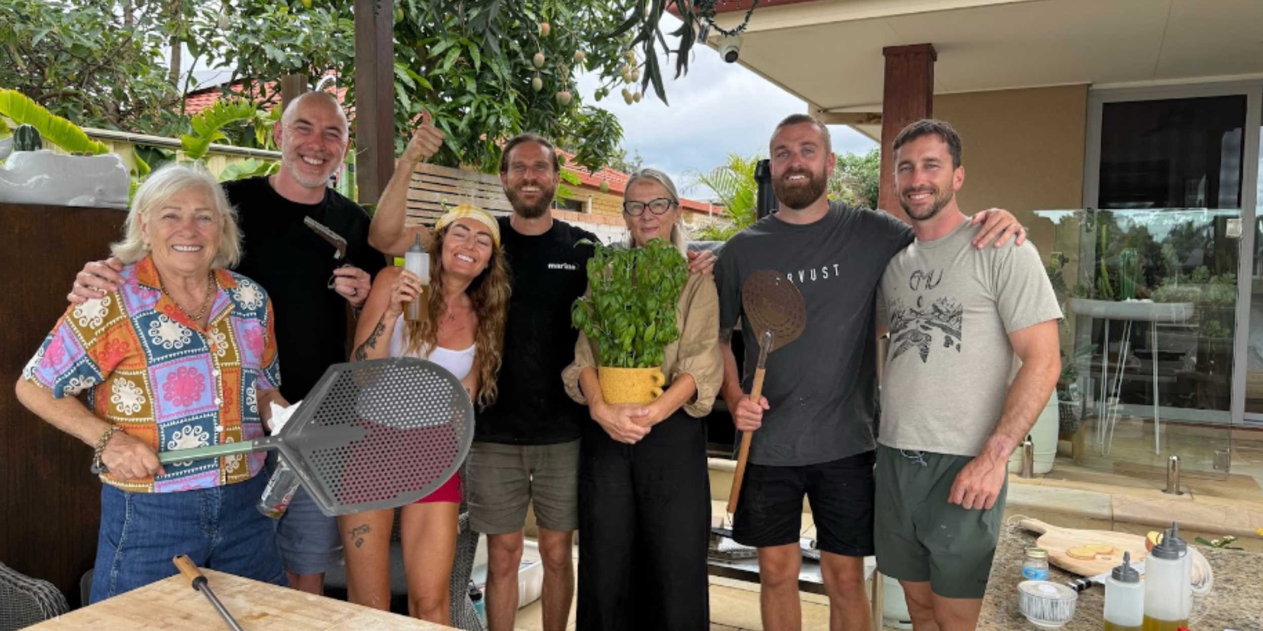 A group of smiling participants poses with dough tools, fresh basil, and olive oil after a Marino & The Dough private masterclass, set outdoors under lush greenery on the Gold Coast.