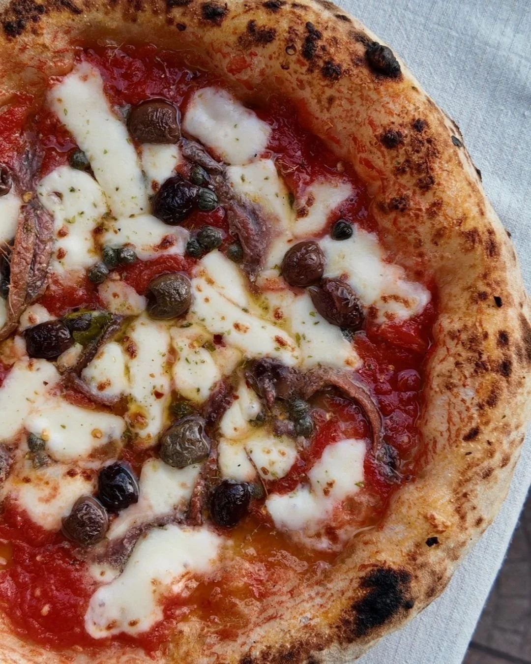 Close up of a Napoli pizza topped with tomato sauce, melted mozzarella, anchovies, black olives and capers on a blistered, wood fired crust.