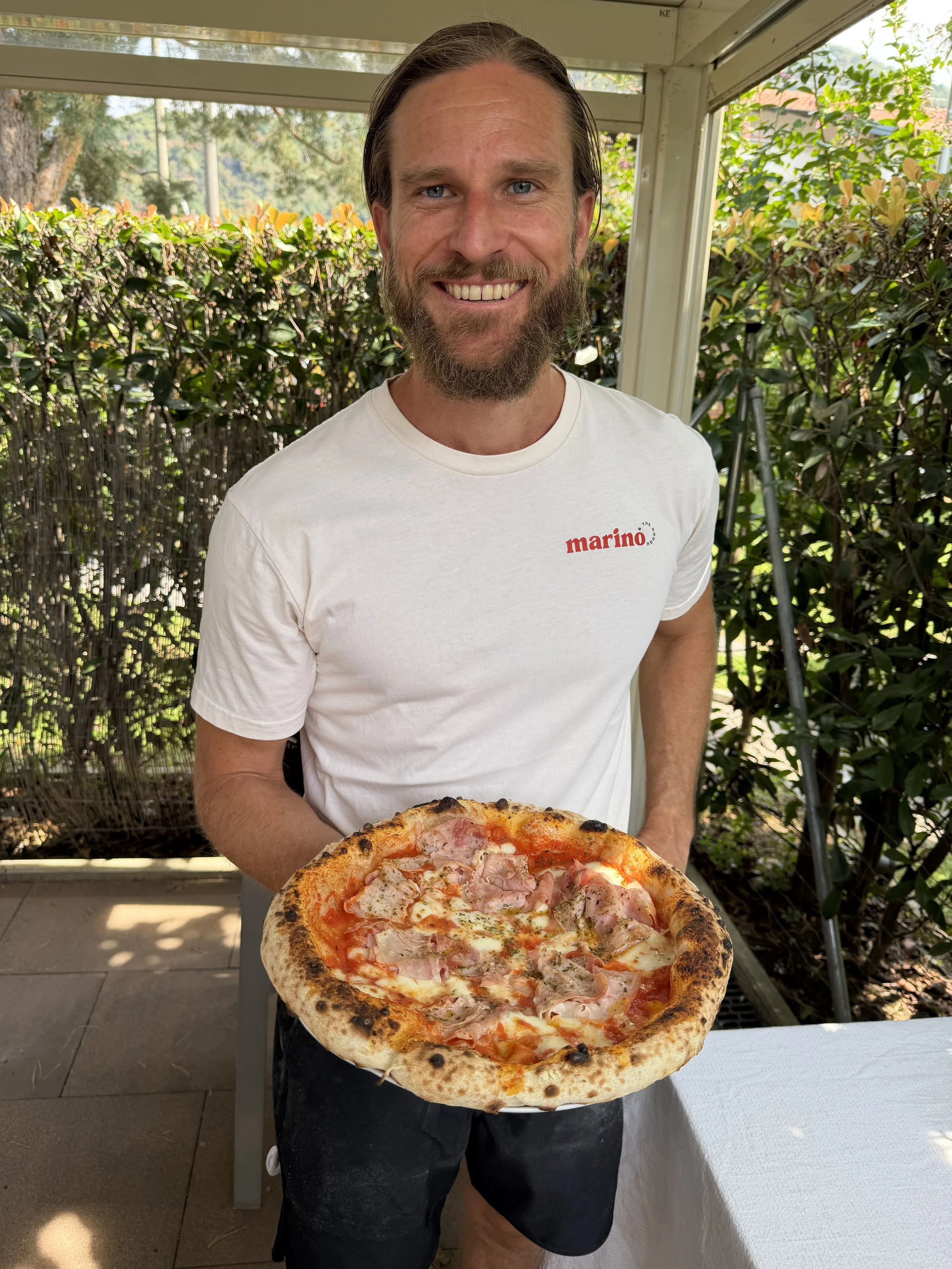 Luke Marino smiling and holding a freshly cooked Neapolitan pizza with a blistered crust, tomato, cheese and ham.