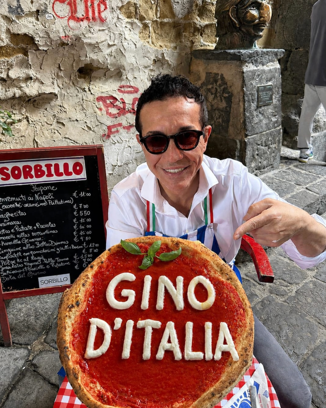 Smiling man in sunglasses and an Italian tricolour apron pointing to a large tomato pizza decorated with mozzarella spelling “Gino d’Italia” outside Sorbillo in Naples.