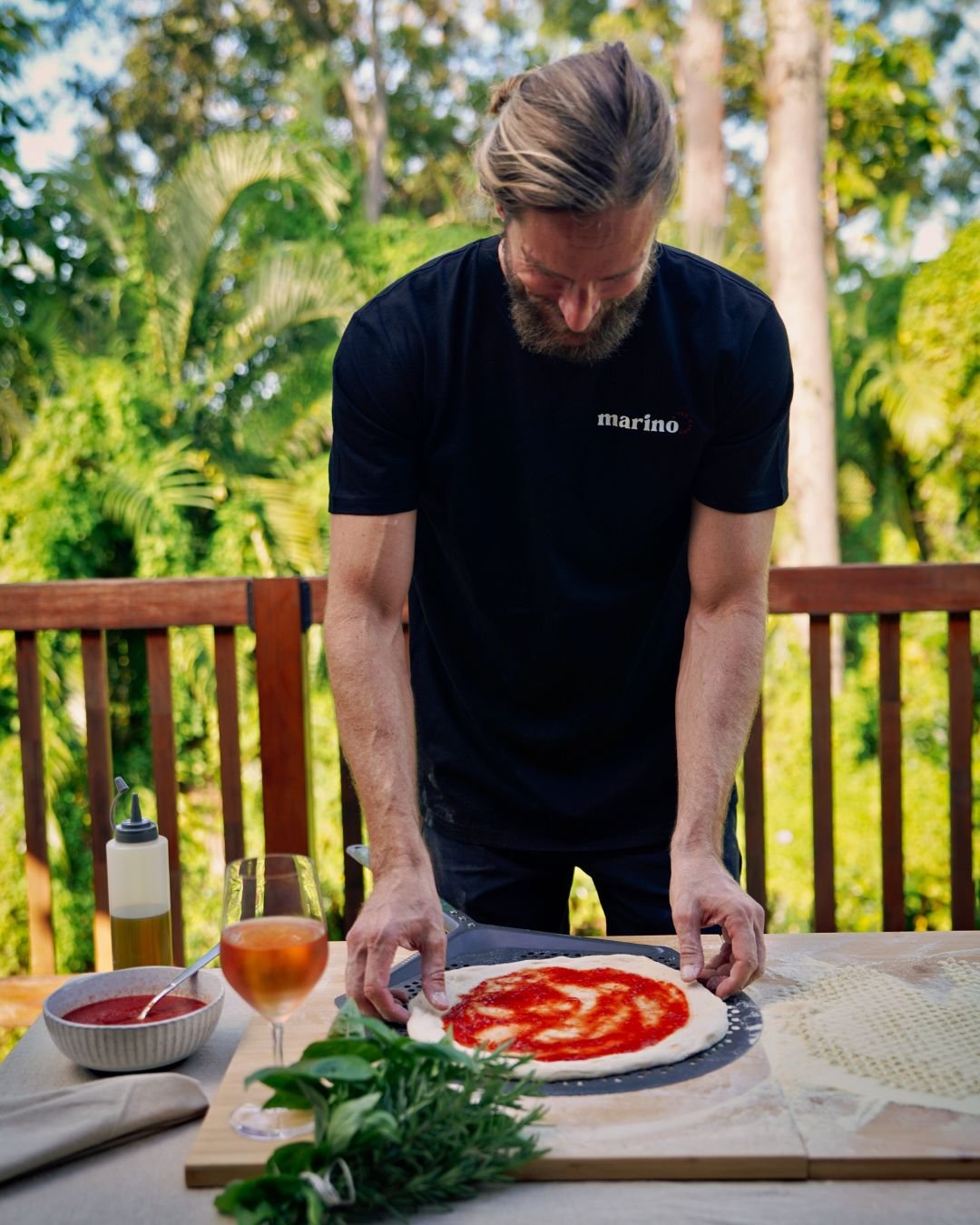 Luke Marino prepares a sauced pizza base on a peel during an outdoor Marino & The Dough masterclass, surrounded by herbs, olive oil, and aperitivo under the trees.