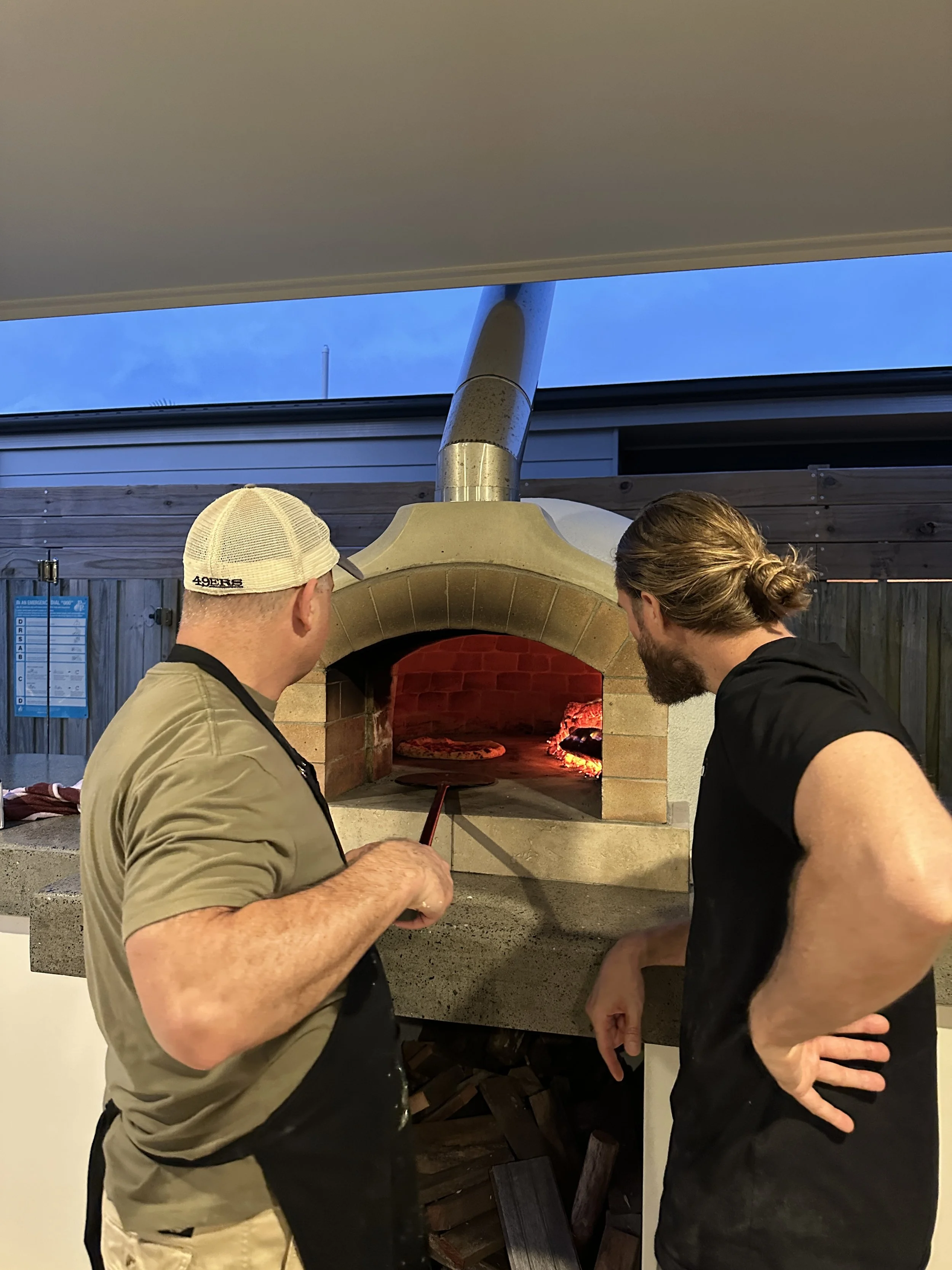 Two guests watching a Neapolitan pizza bake inside a glowing wood-fired oven at a Marino & The Dough masterclass
