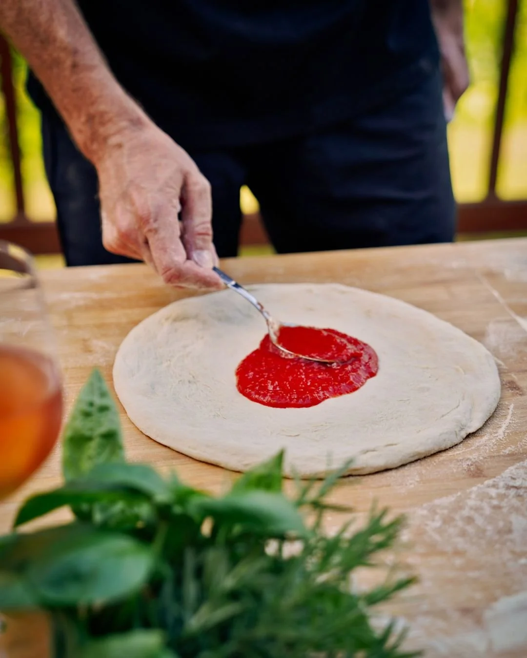 A spoon gently spreads rich tomato sauce over freshly shaped pizza dough during a Marino & The Dough outdoor masterclass, with herbs and an aperitif in soft focus.