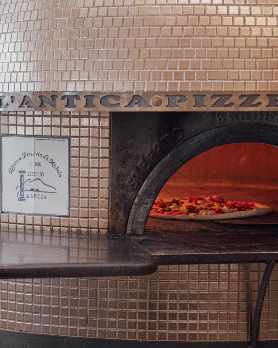 Pizza baking inside the wood fired oven at L’Antica Pizzeria, framed by tiled walls and the oven entrance