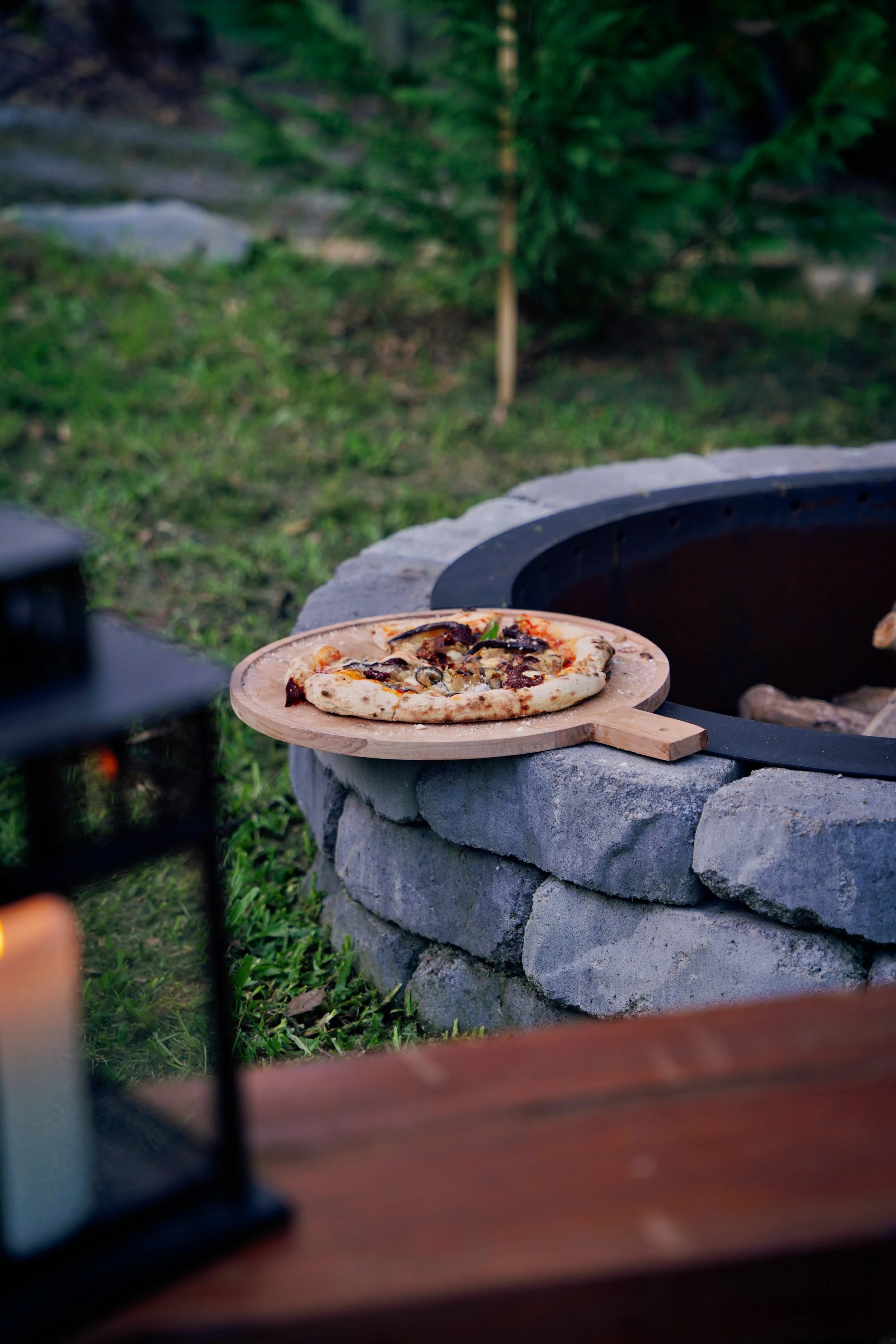 A wood-fired pizza on a wooden peel rests atop a stone firepit in a backyard setting.