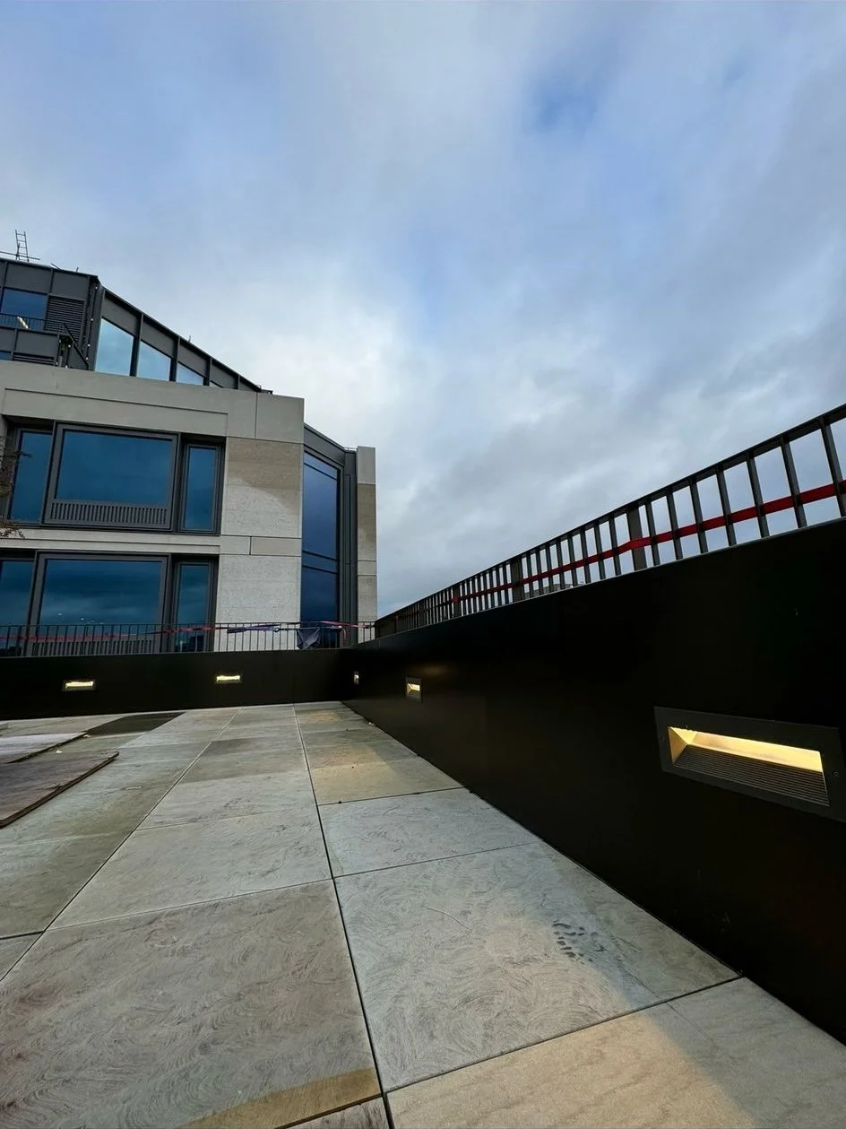 Modern rooftop terrace with gray tiles, an adjacent building with large windows, and ambient lighting on a cloudy day.