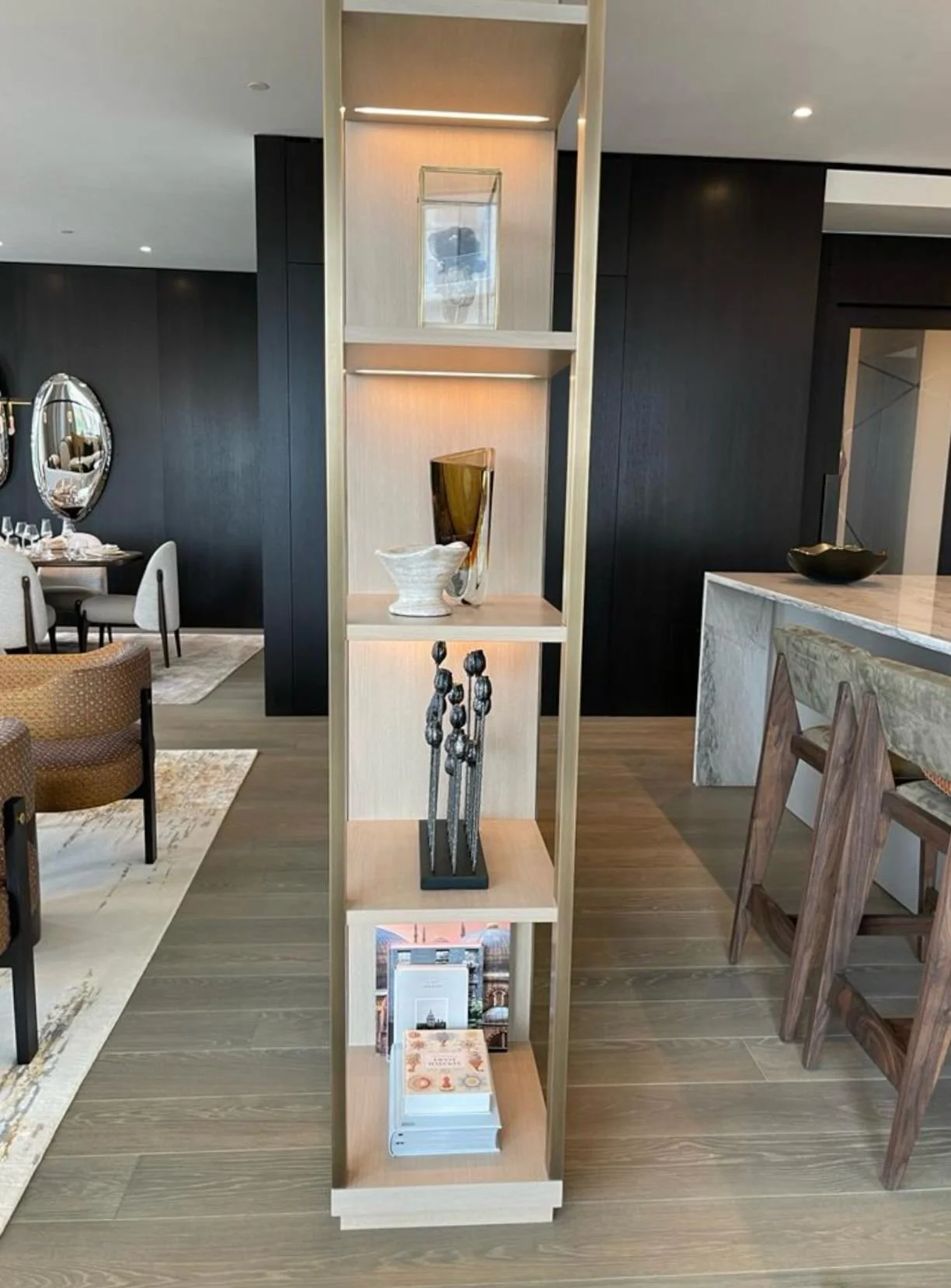 Modern room with open shelves adorned with decorative items, including a clear glass square container, white ceramic piece, abstract sculpture, and stacked books. The setting includes elegant dining and seating areas in a contemporary space.