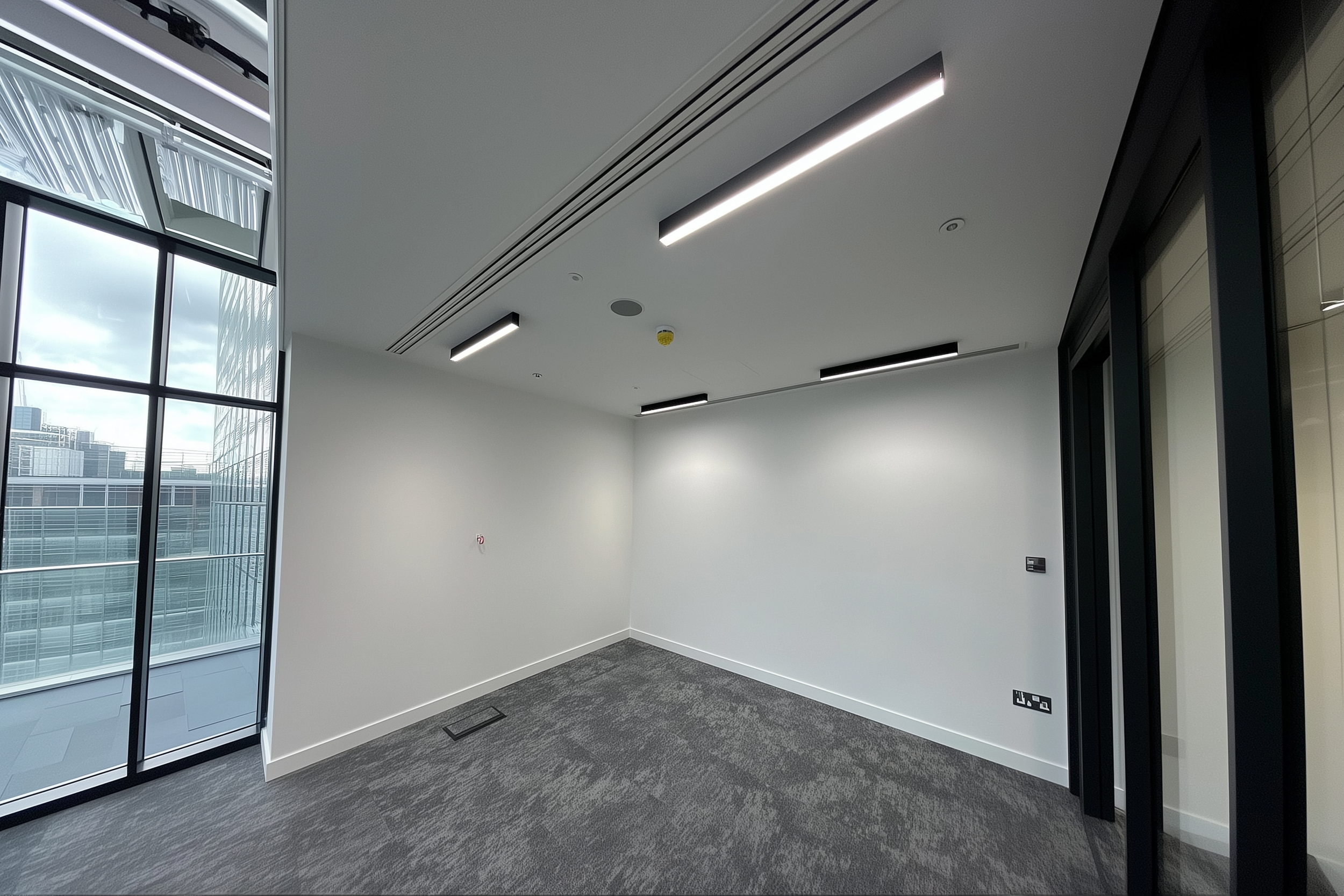 Empty modern office space with carpet flooring, white walls, and ceiling lights. Floor-to-ceiling windows provide a view of urban buildings. One wall has a control panel and power outlets.