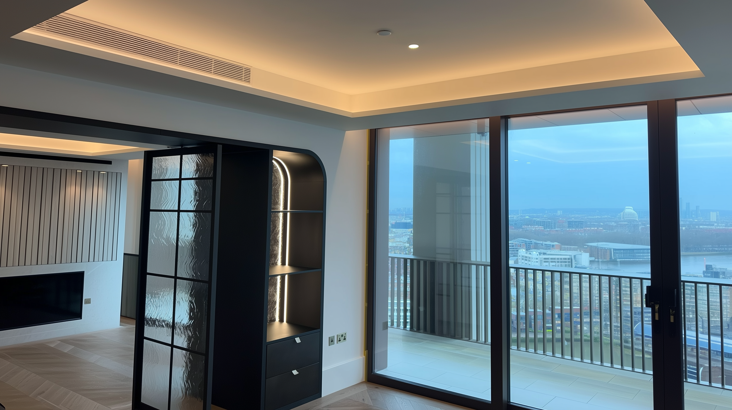 Modern living space with large windows, wooden flooring, built-in shelving, and a view of a cityscape and river.