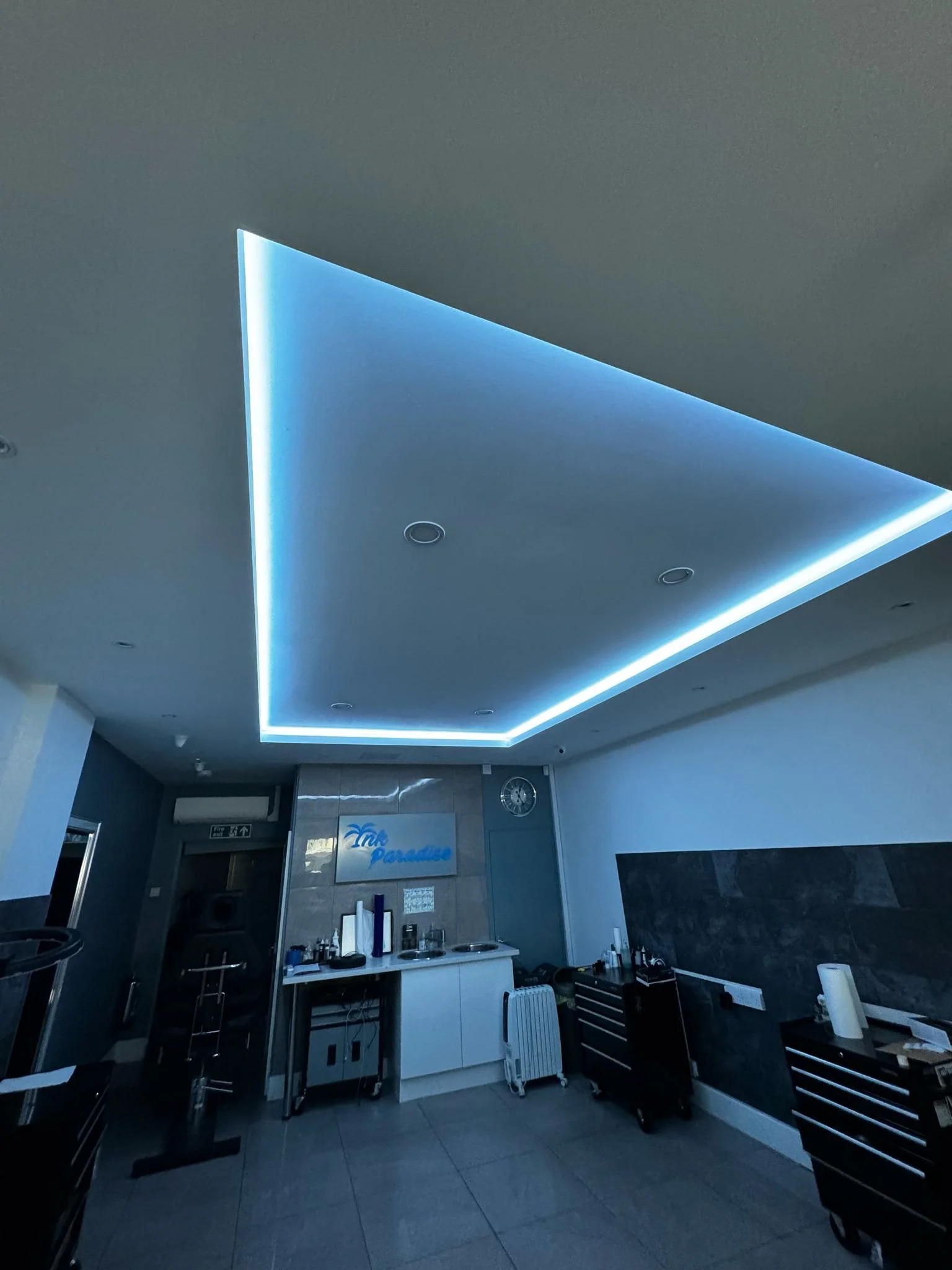 Room with modern ceiling lights, blue LED strip lighting, a wall clock, and a tiled wall with a sign reading "Paradise." The space includes storage cabinets and equipment.