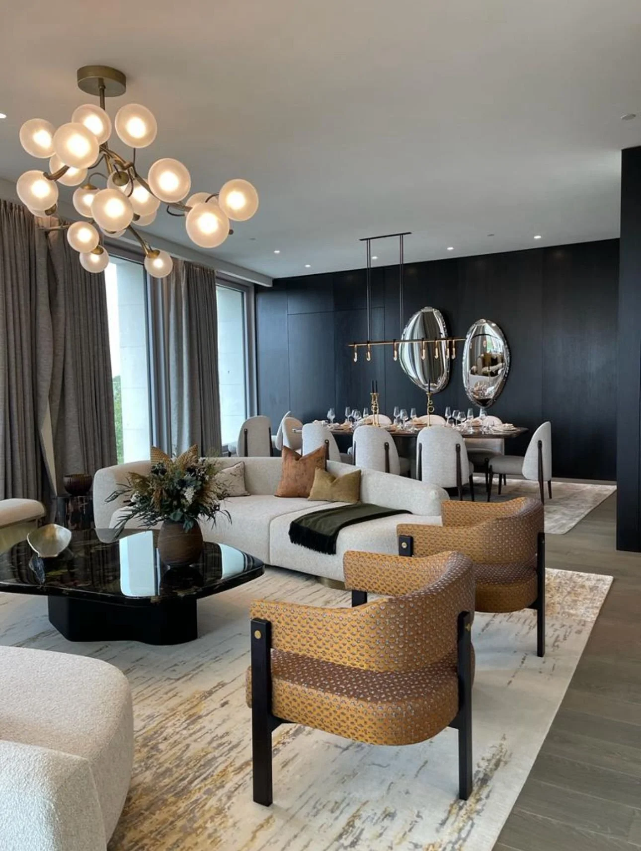 Modern living and dining room with stylish furniture and decor, featuring a contemporary light fixture, plush seating, a dark glossy coffee table, a carpet, and an elegant dining area with mirrored decor.