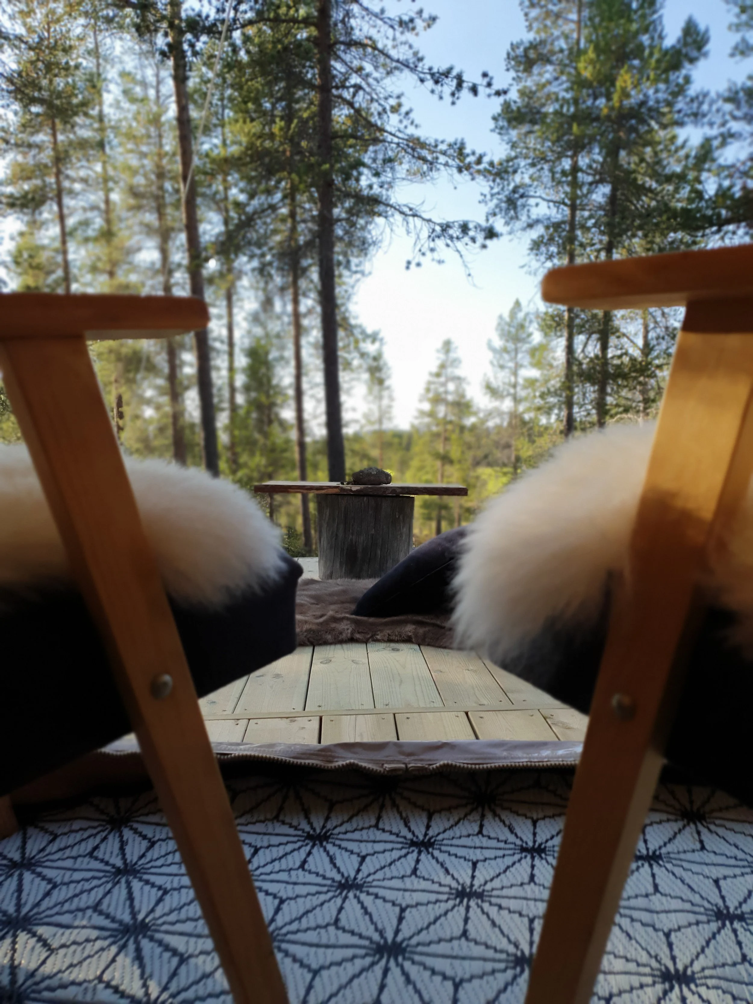 Sitzgruppe im Freien mit zwei Holzstühlen mit weißen Fellkissen vor einem Tisch aus einem Baumstumpf, umgeben von einem Wald aus hohen Bäumen.