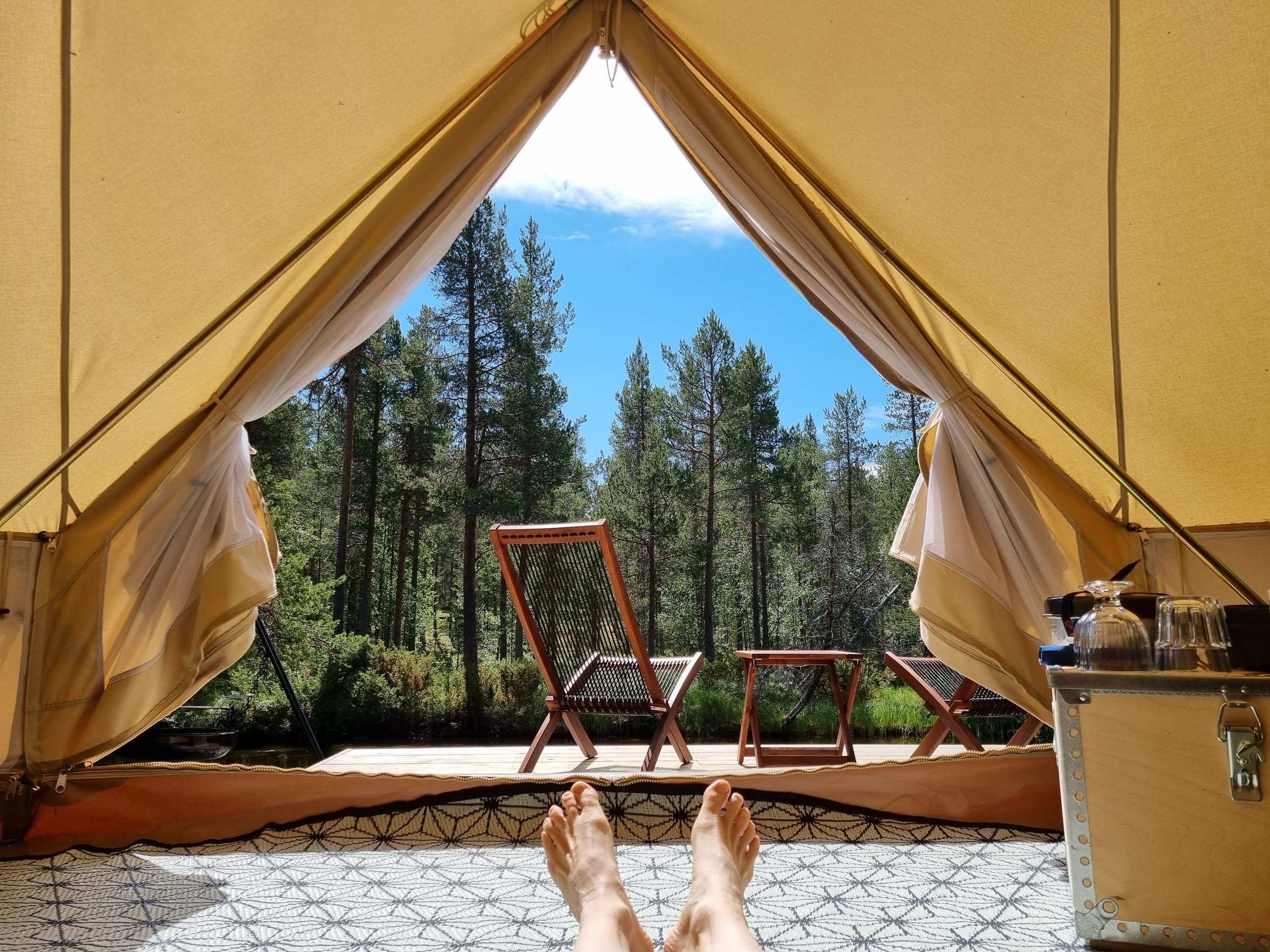 Utsikt fra innsiden av et glampingtelt som viser føtter, campingstoler, et lite bord og skogslandskap.