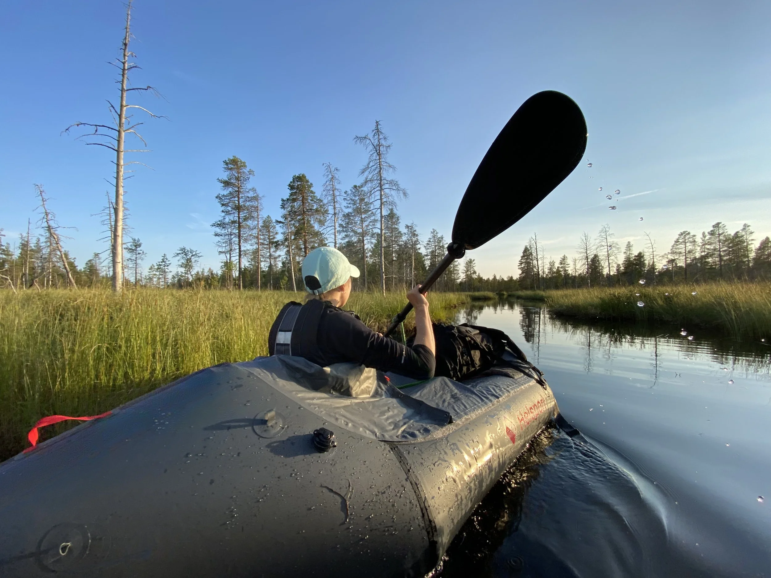 Person kayaking on a calm river surrounded by tall trees and green grass during sunset.