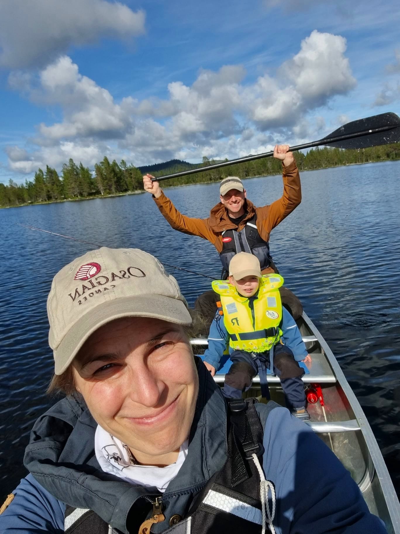 A family of three canoeing on a lake. A woman in a cap and life jacket is smiling at the camera. Behind her, a child in a bright yellow life jacket is seated, and a man in a cap and jacket is with a paddle raised in celebration. The lake is surrounde