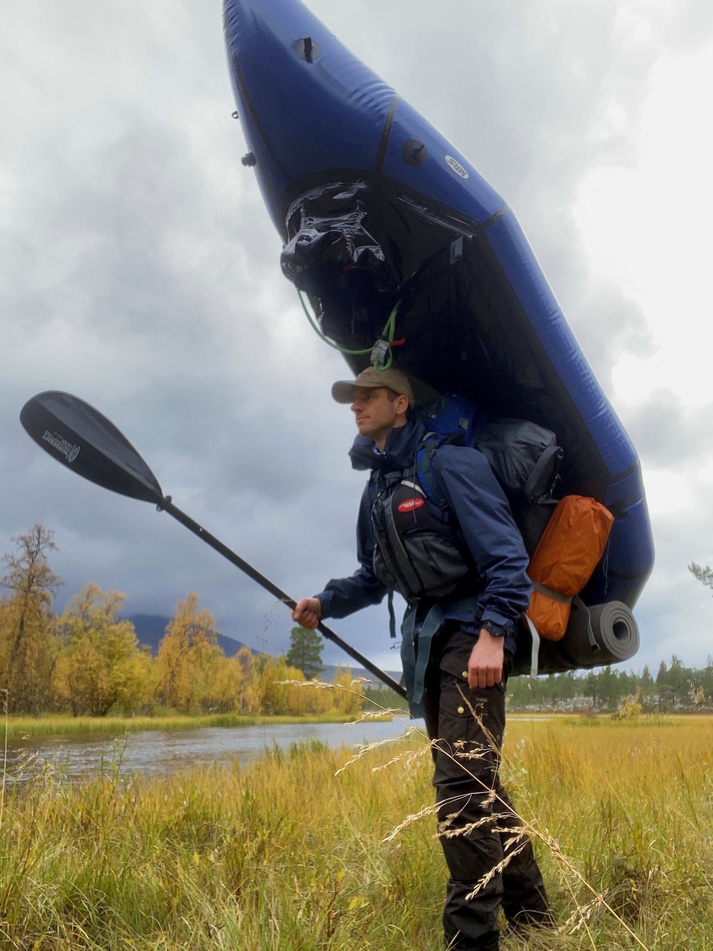 En person som vandrer i et gresskledd område nær en elv, bærer en oppblåsbar packraft og en padle. De har på seg utendørsklær og en ryggsekk med campingutstyr festet til.