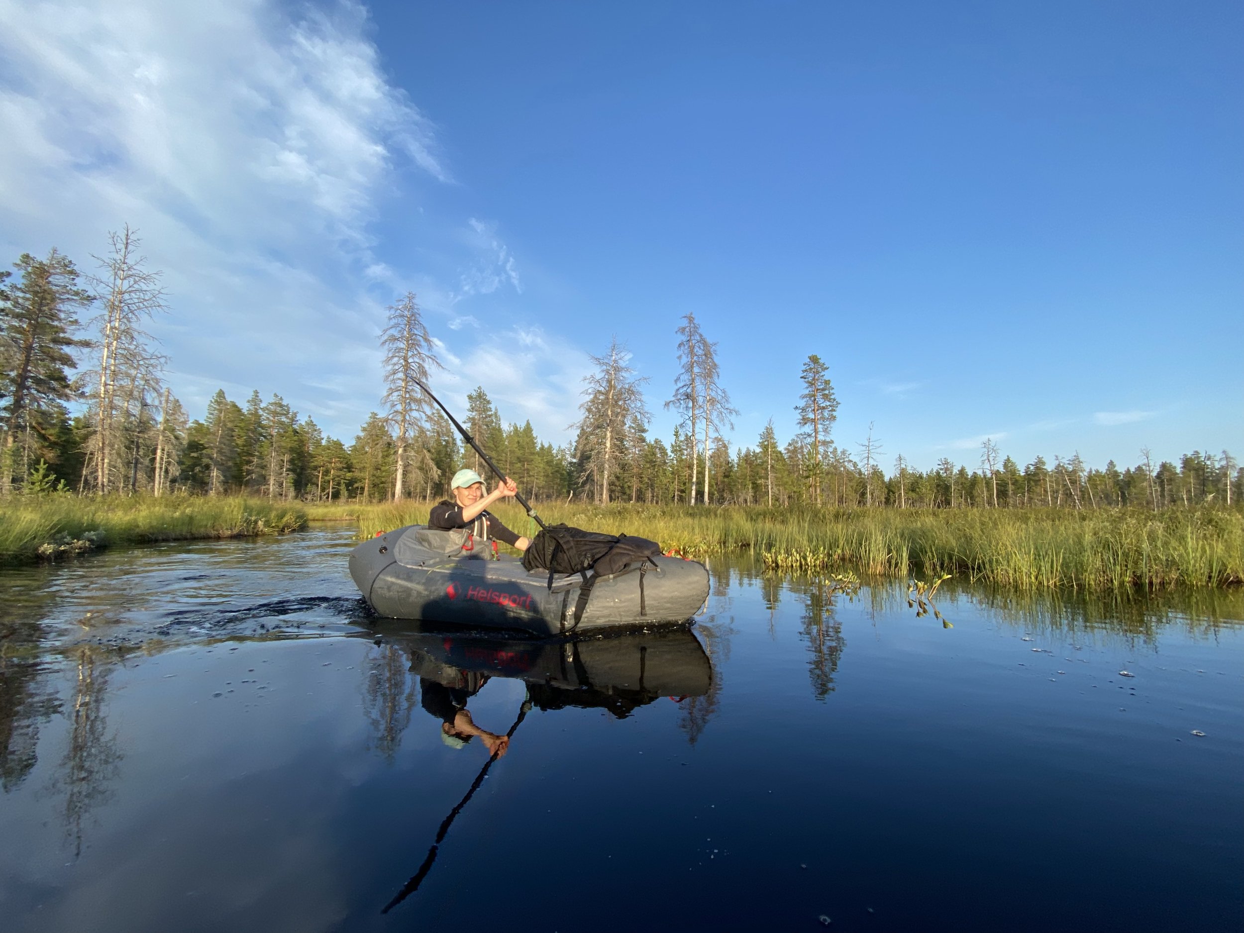 Person som padler en oppblåsbar båt packraft i en rolig innsjø omgitt av skog og høyt gress under en klar blå himmel.