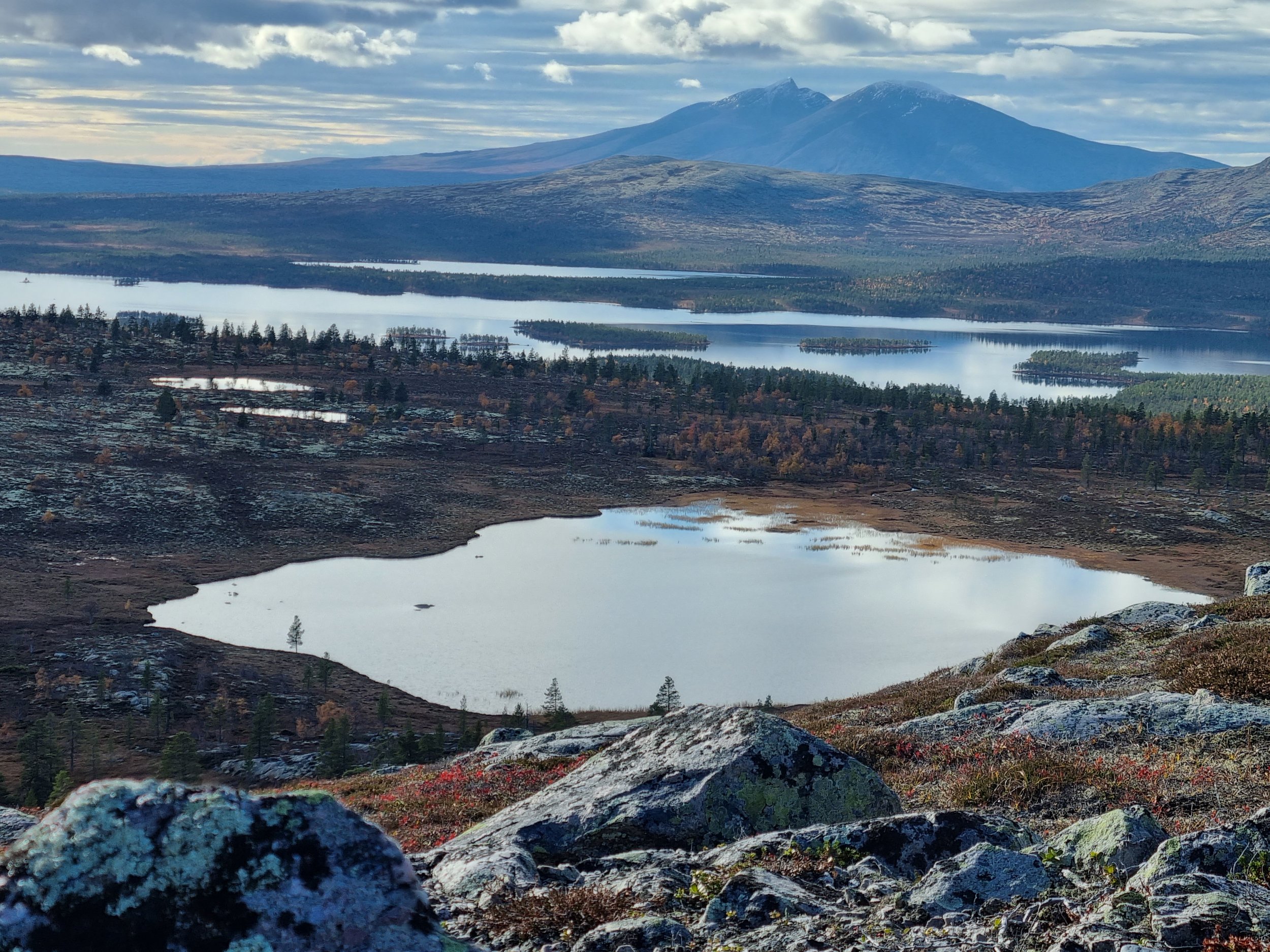 Scenic landscape with mountains, lakes, and a rocky foreground, under a cloudy sky.