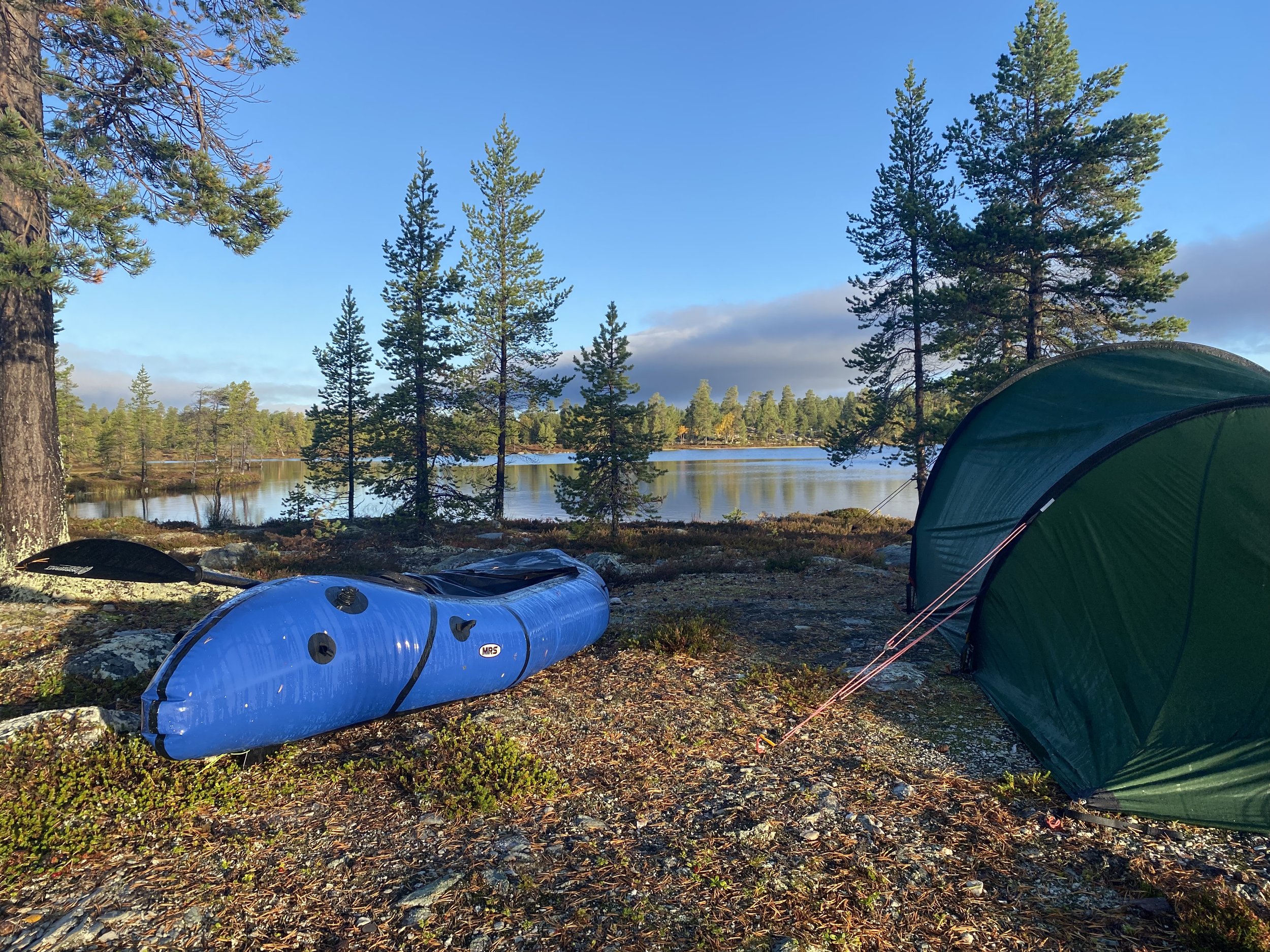 Campingscene med en blå oppblåsbar bil packraft , et grønt telt, furutrær og en rolig innsjø under en klar himmel.