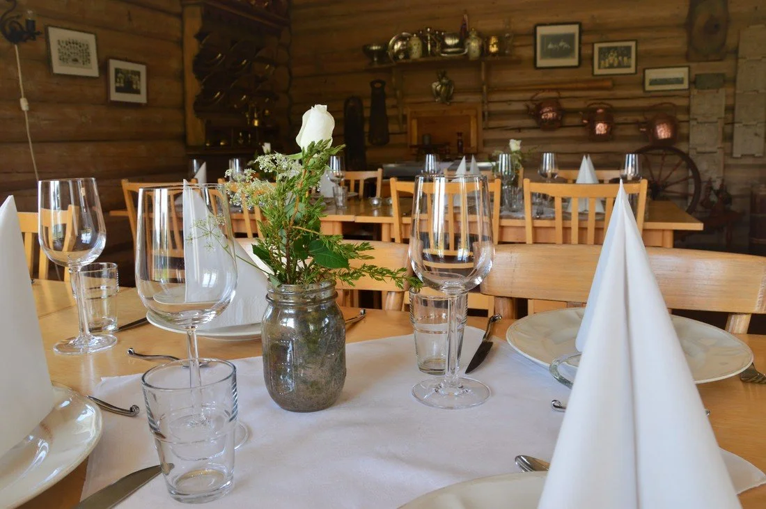 Tables set for a meal in a rustic wood-paneled restaurant with wine glasses, white napkins, plates, and a small vase with a white flower and greenery.